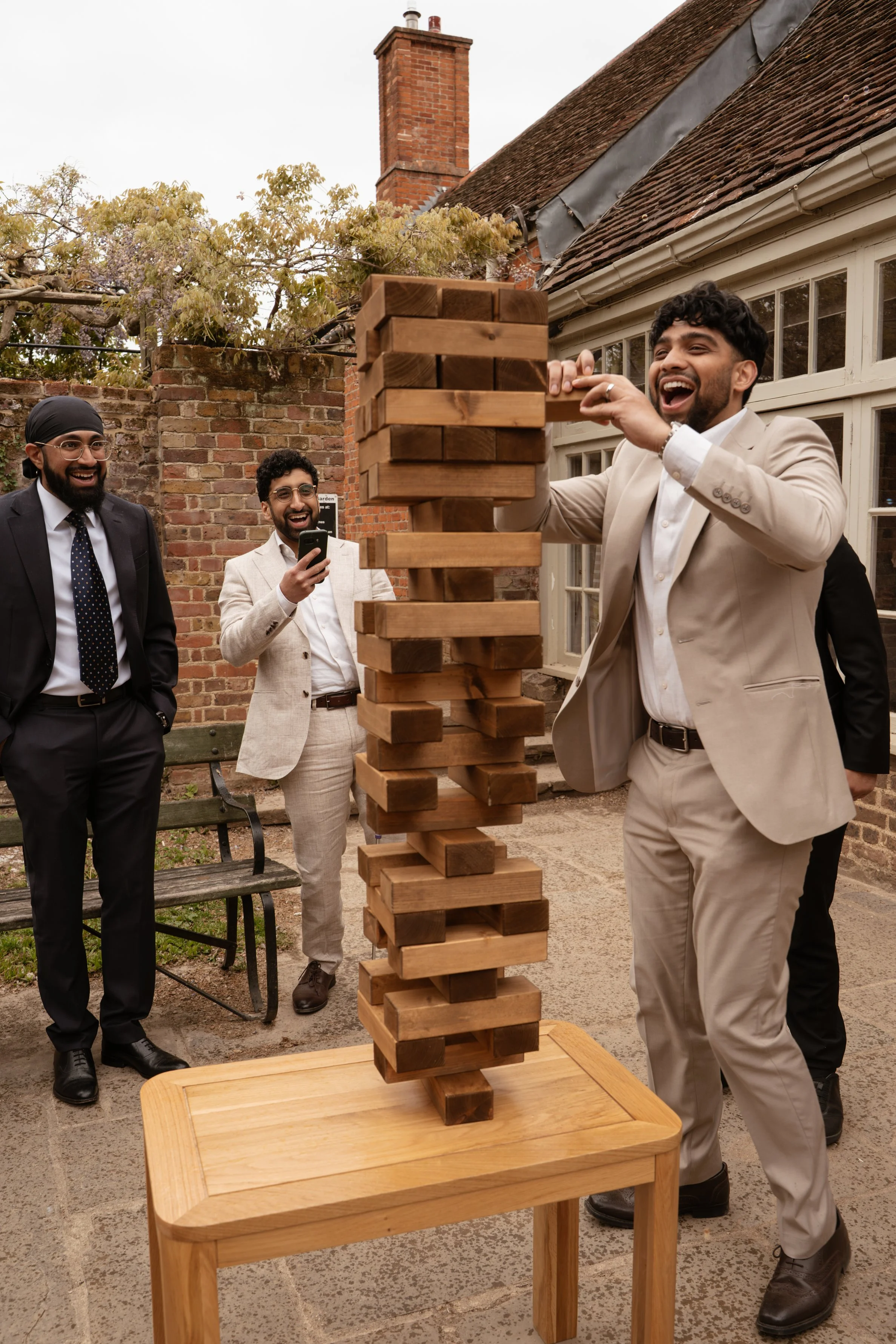 Group of men playing giant outdoor Jenga game in a yard with brick and wooden houses, some dressed in suits, one smiling while playing, others watching and smiling, one taking a photo.