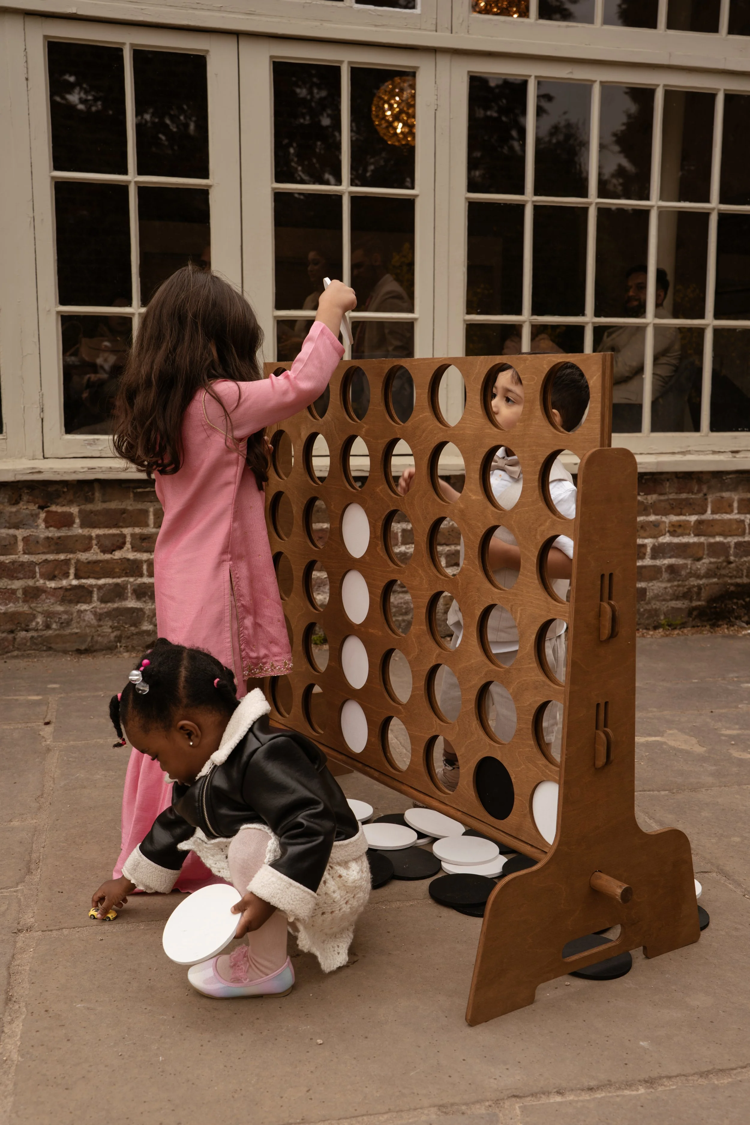 Children playing a large connect four game outdoors near a brick building and large window, with some children and adults visible through the window.