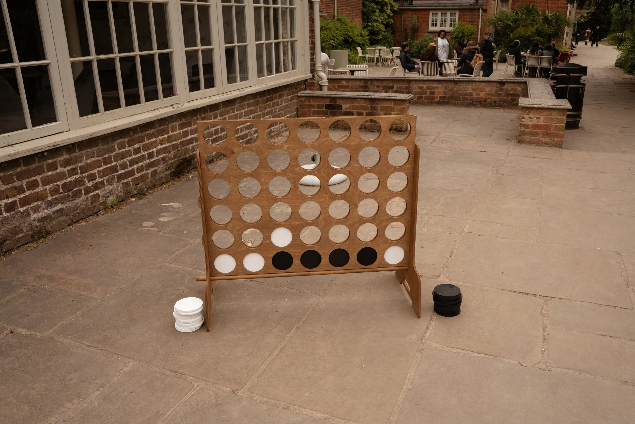 An outdoor setting with a large wooden Connect Four game set up on a pavement. There are stacks of white and black game pieces near the game. In the background, people are sitting and standing at tables outside, at a wedding.