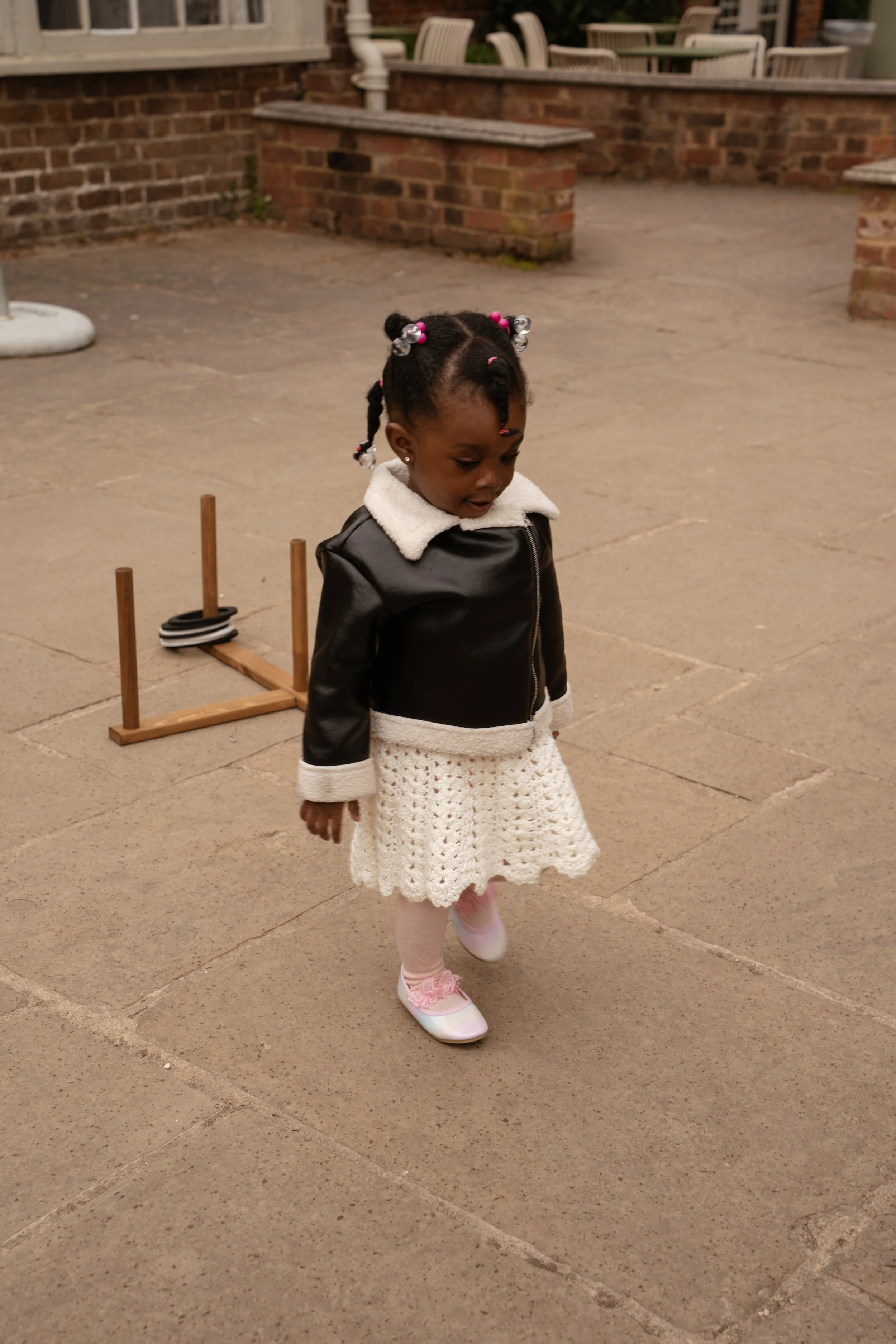 A young girl with braided hair decorated with beads, wearing a black and white jacket, a white knitted skirt, pink tights, and pink shoes, walking on a concrete surface outside near a brick wall with a wooden game set in the background.