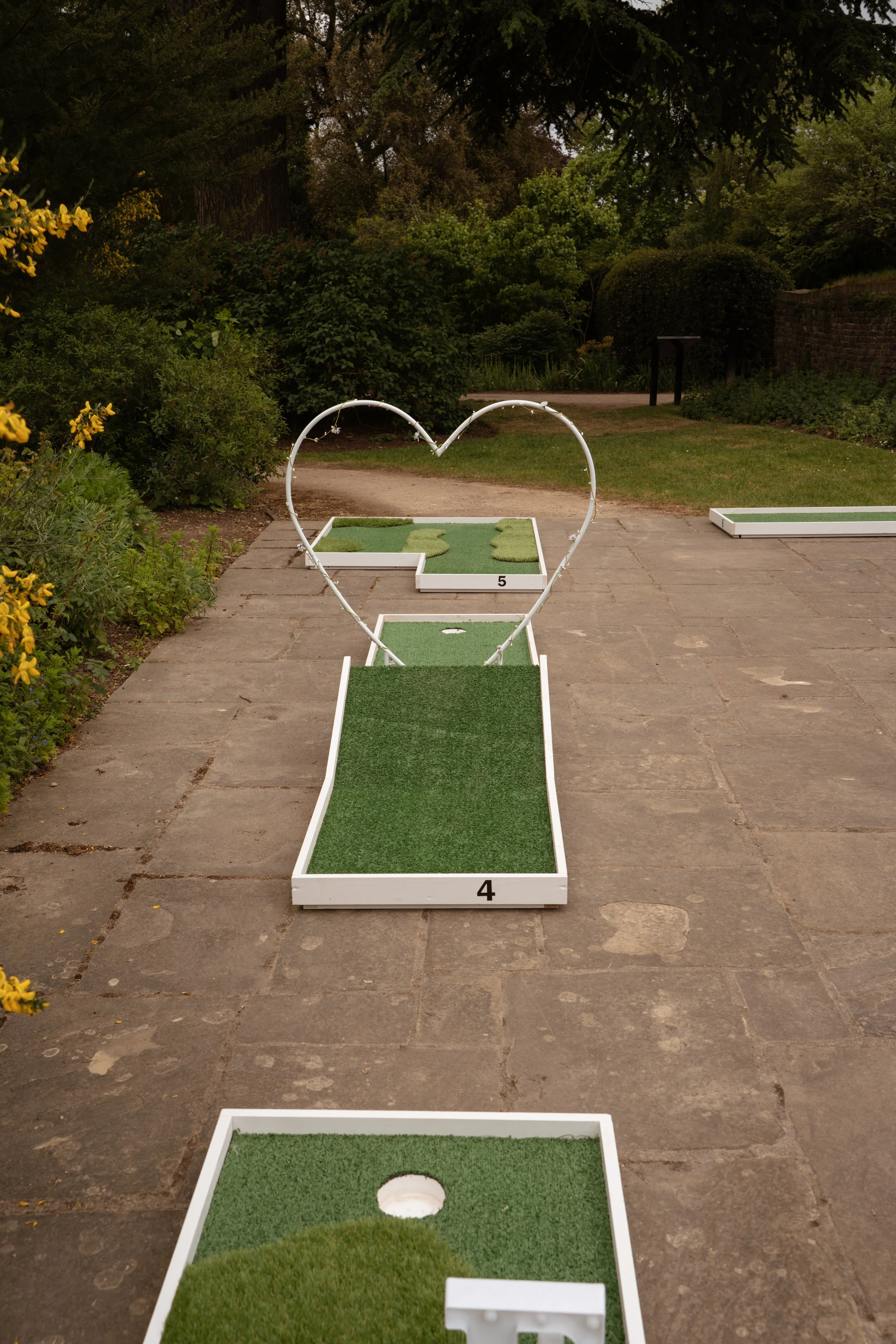 Miniature golf course with numbered holes and a heart-shaped decoration, set in a garden or park.