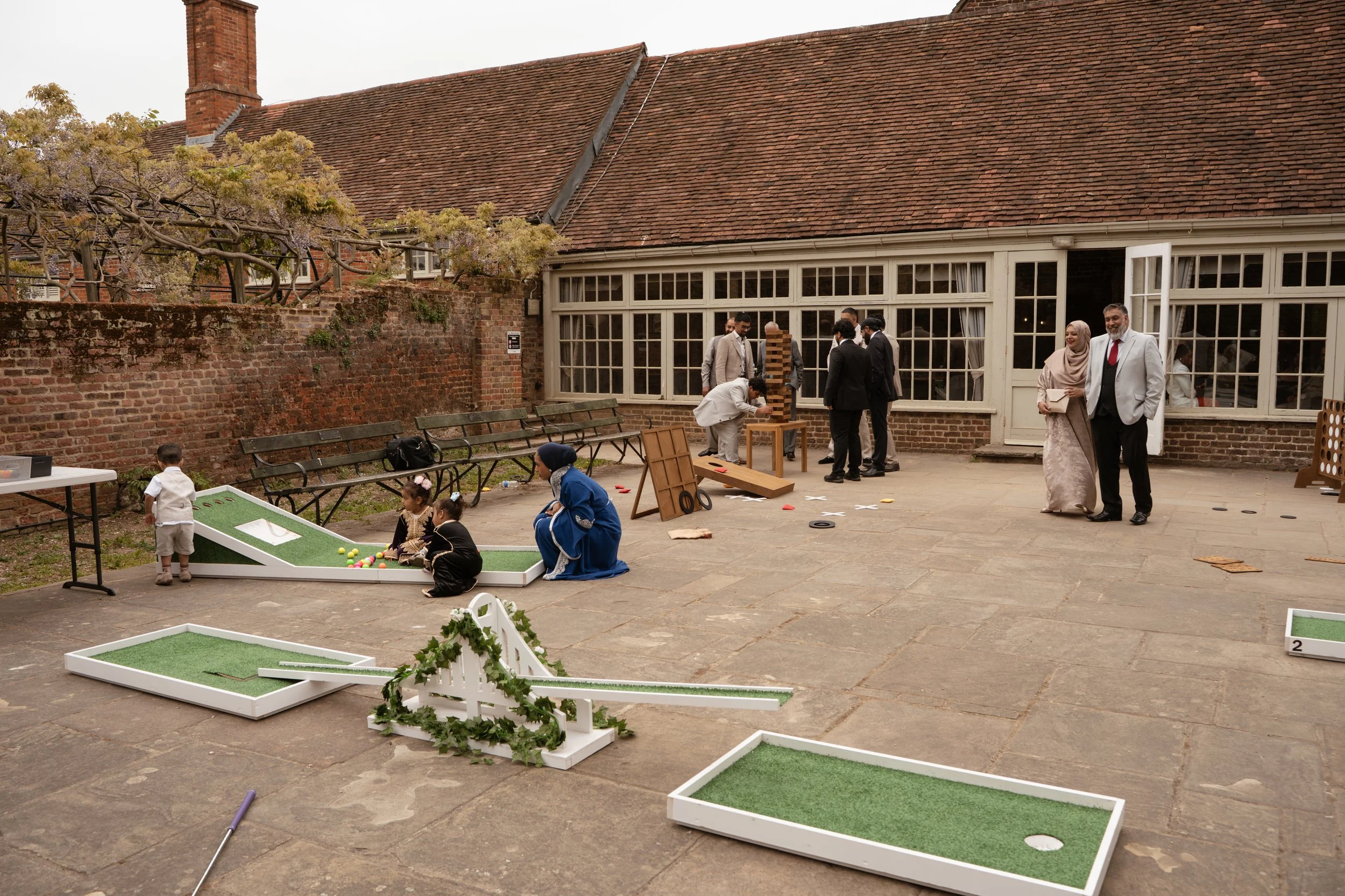 People playing mini-golf, socializing, and participating in outdoor activities in a courtyard with benches, trees, and an old brick building with large windows.