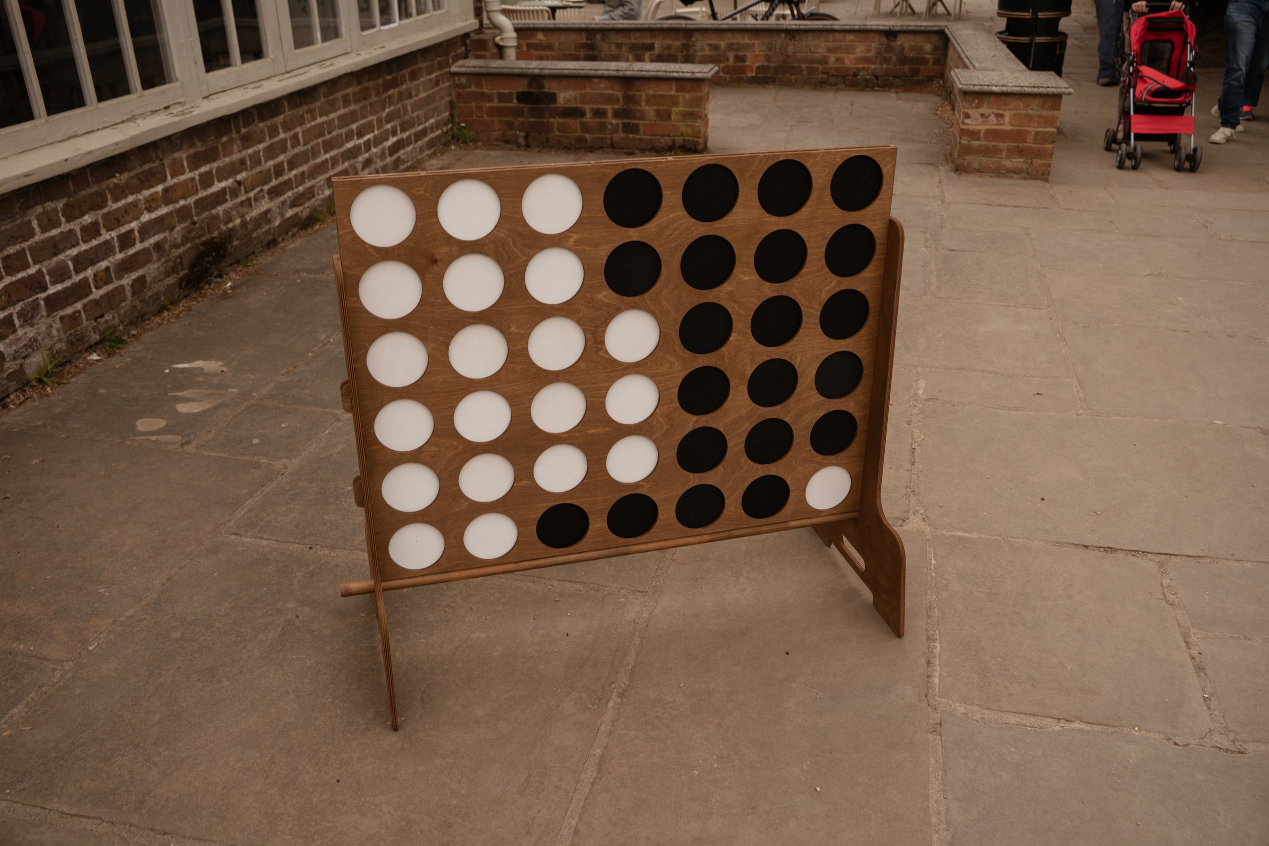 A large wooden Connect Four game board with black and white circular pieces, placed outside on stone pavement near a brick building. In the background, people are visible, including a person with a red stroller.