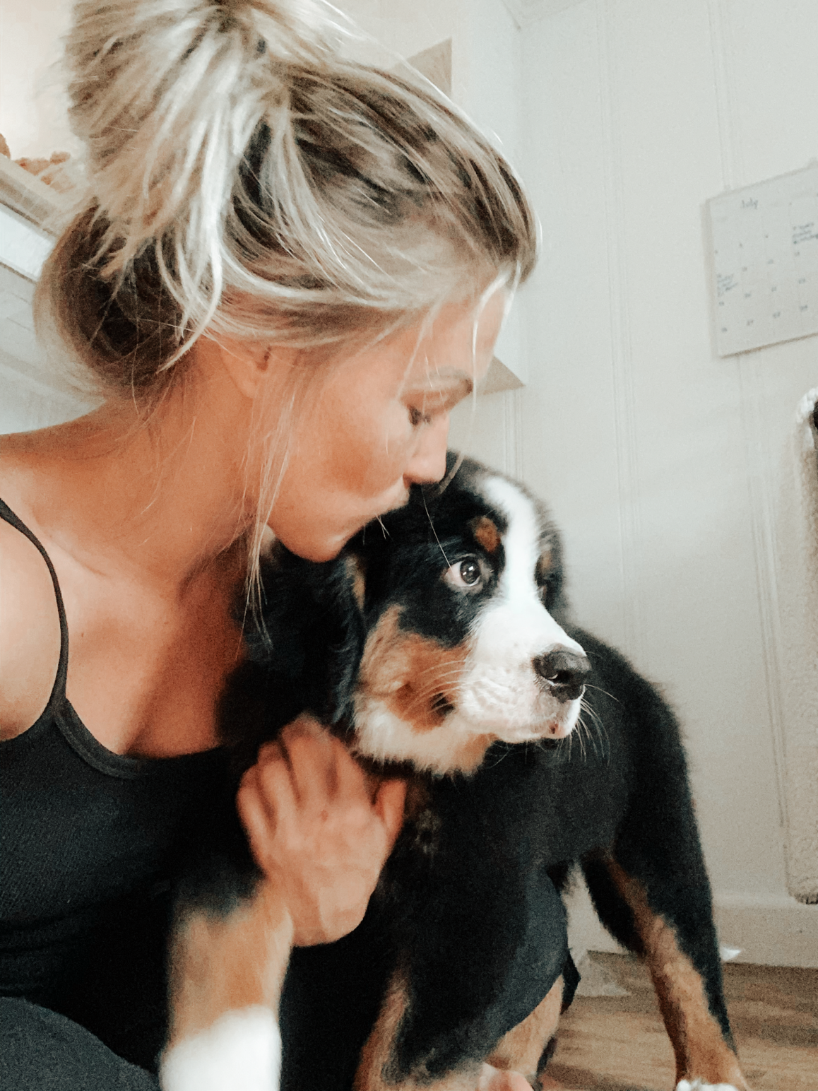 A woman with blonde hair in a bun leaning in to kiss a black, white, and tan Australian Shepherd puppy.