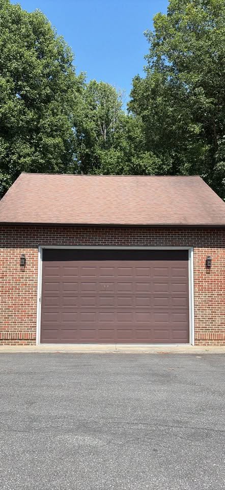 A brick garage with a brown door, two outdoor wall lights, and a shingled roof in front of trees and a blue sky.