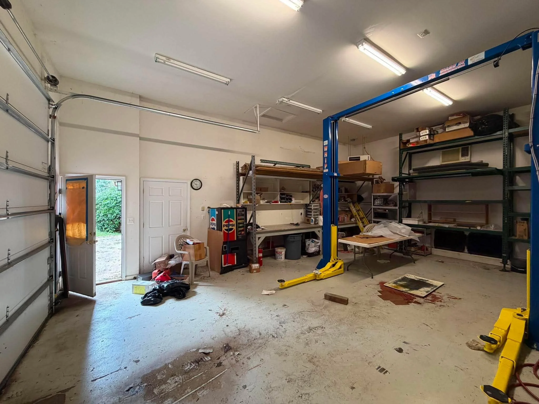 A garage with a car lift, shelves, and various items. The floor has oil or grease stains and some debris. Thegarage door is open, showing trees outside.