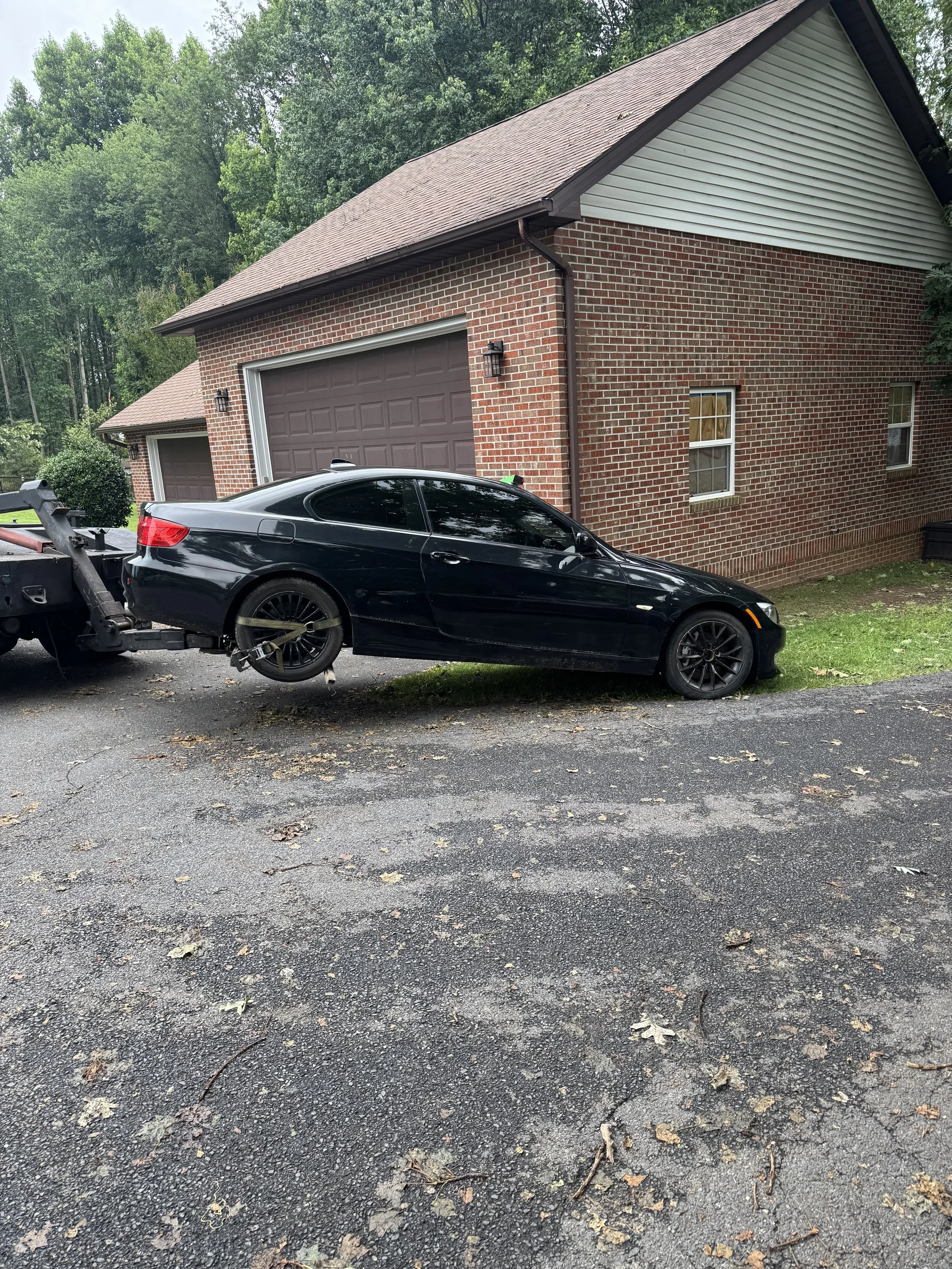 A black sports car parked in front of a brick house with a garage, on an asphalt driveway surrounded by trees and fallen leaves.