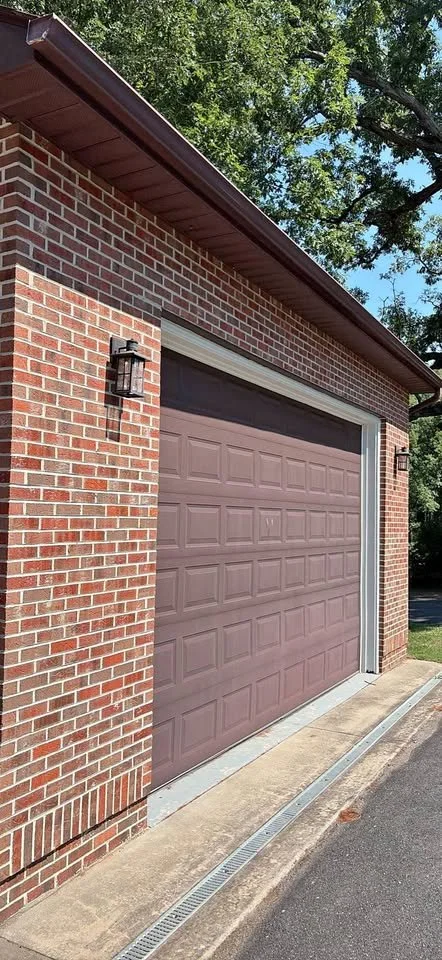 A residential garage with a brick exterior, a closed purple garage door, two black outdoor wall lanterns, a concrete driveway with drainage grate, and trees in the background.