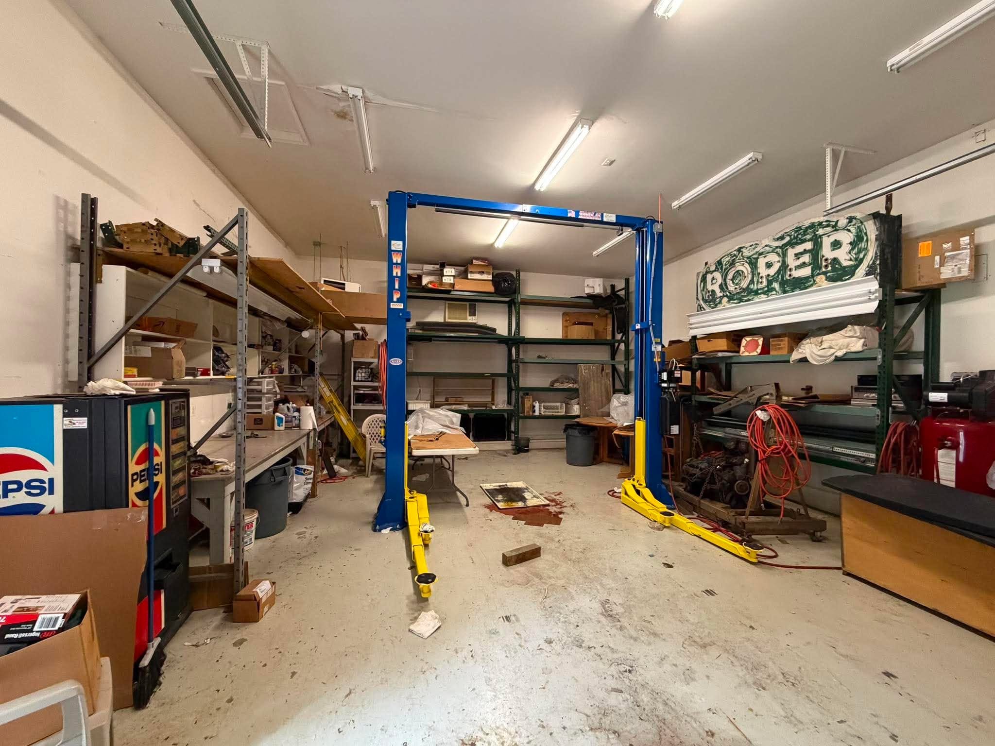 A cluttered garage storage space with shelves filled with various items including boxes, tools, and bottles. A blue hydraulic lift is centered in the room, and there are fluorescent lights on the ceiling. The floor has some stains and scattered objects.