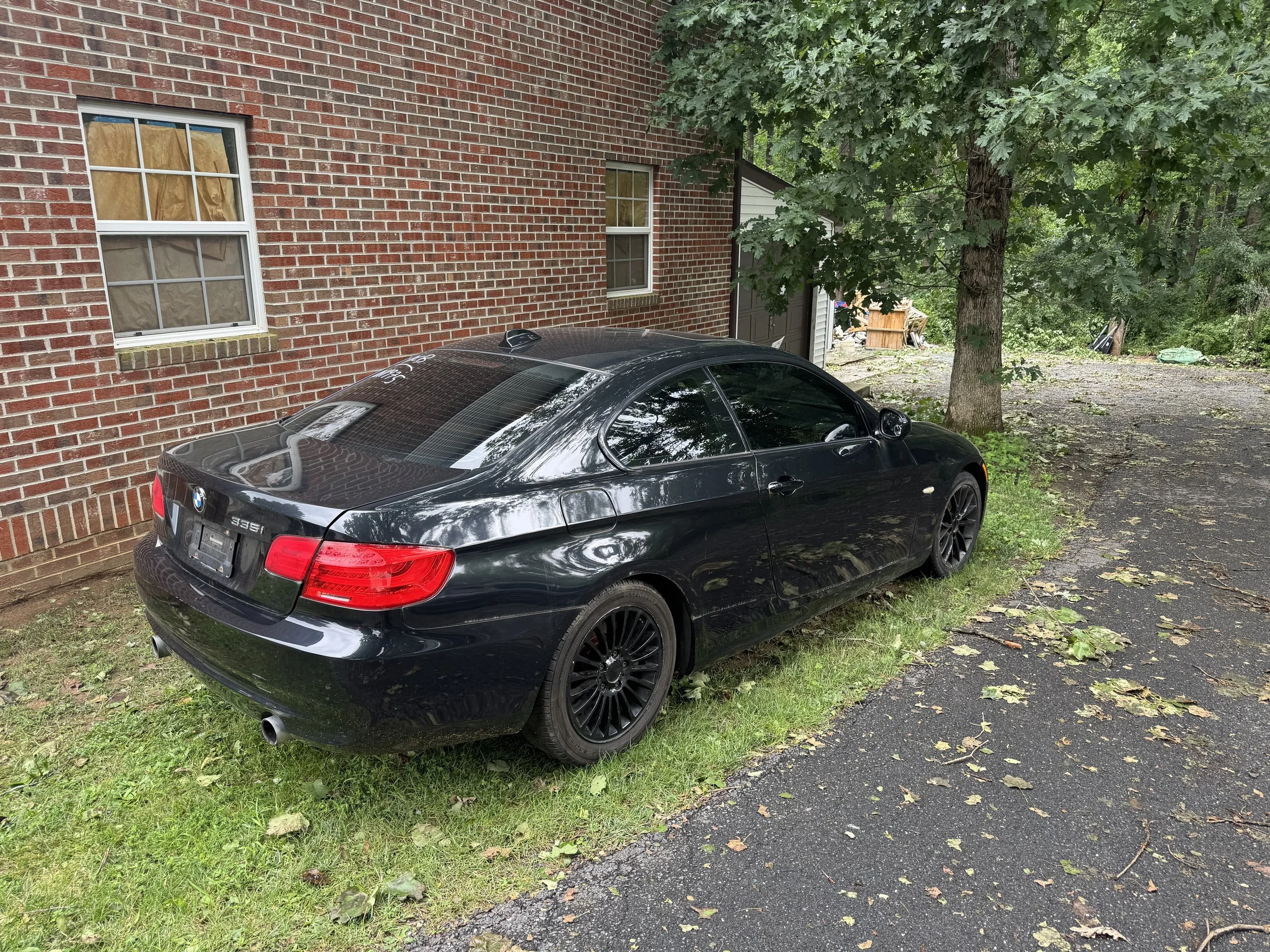Black BMW car parked on a patch of grass and asphalt next to a brick building with two windows, a large tree, and some outdoor clutter in the background.