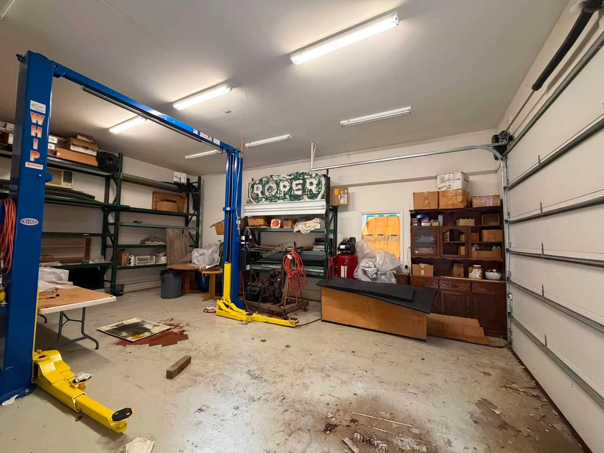 A cluttered garage with shelves holding various items, a sign that reads 'Roper', and a blue vehicle lift. The floor has dirt and debris, and there are fluorescent lights on the ceiling.