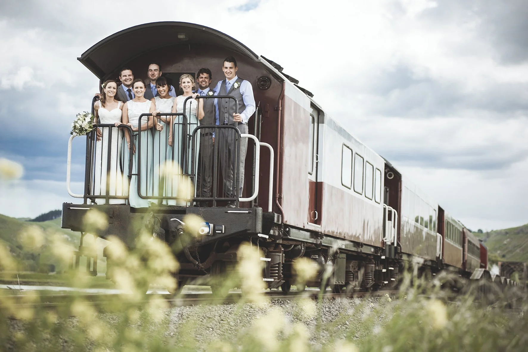 A group of wedding guests, including women in white dresses and men in suits, smiling and standing on the caboose of a train in a rural landscape with green hills and cloudy sky.