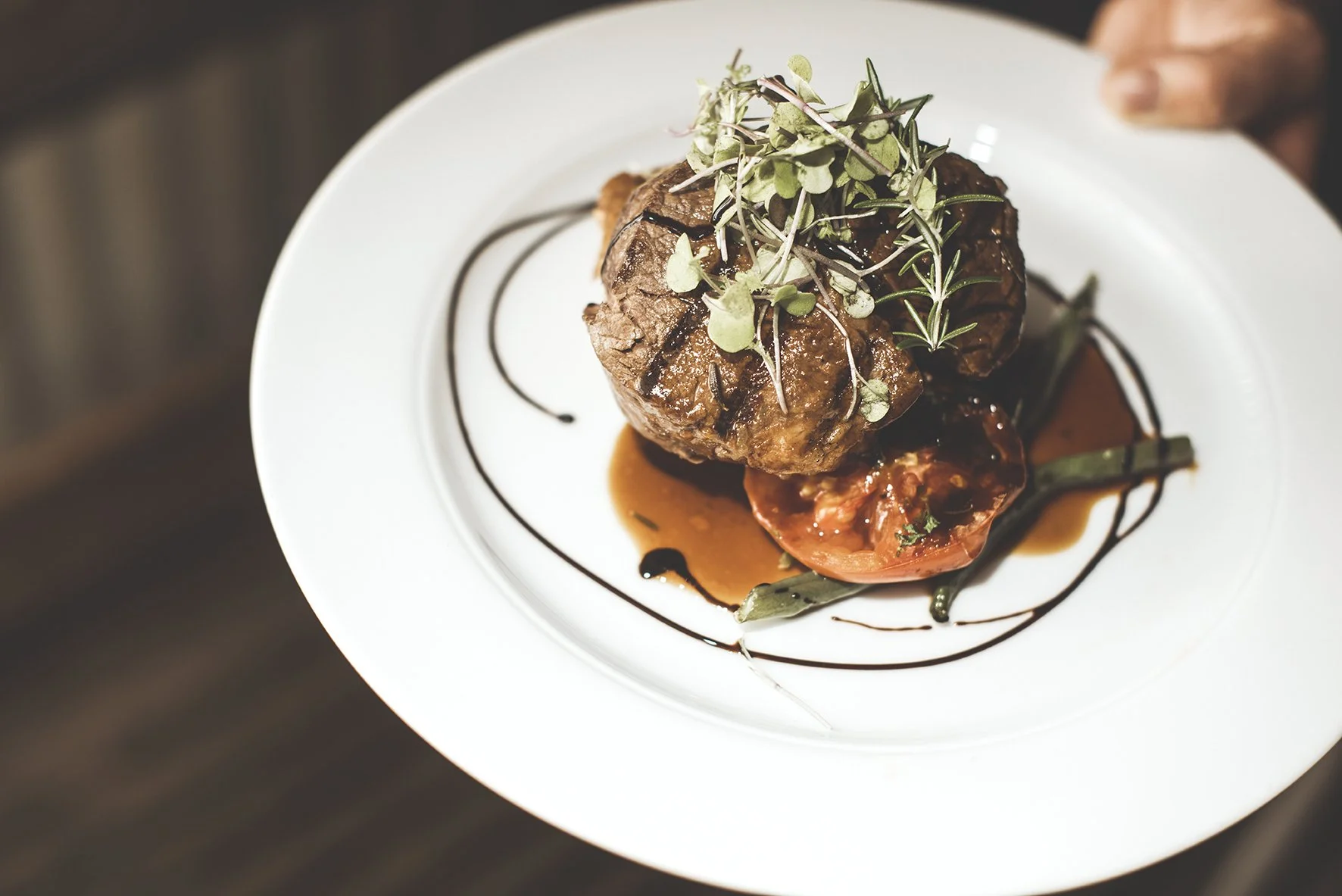A white plate with grilled meat, roasted tomato, green beans, and microgreens, drizzled with balsamic glaze.