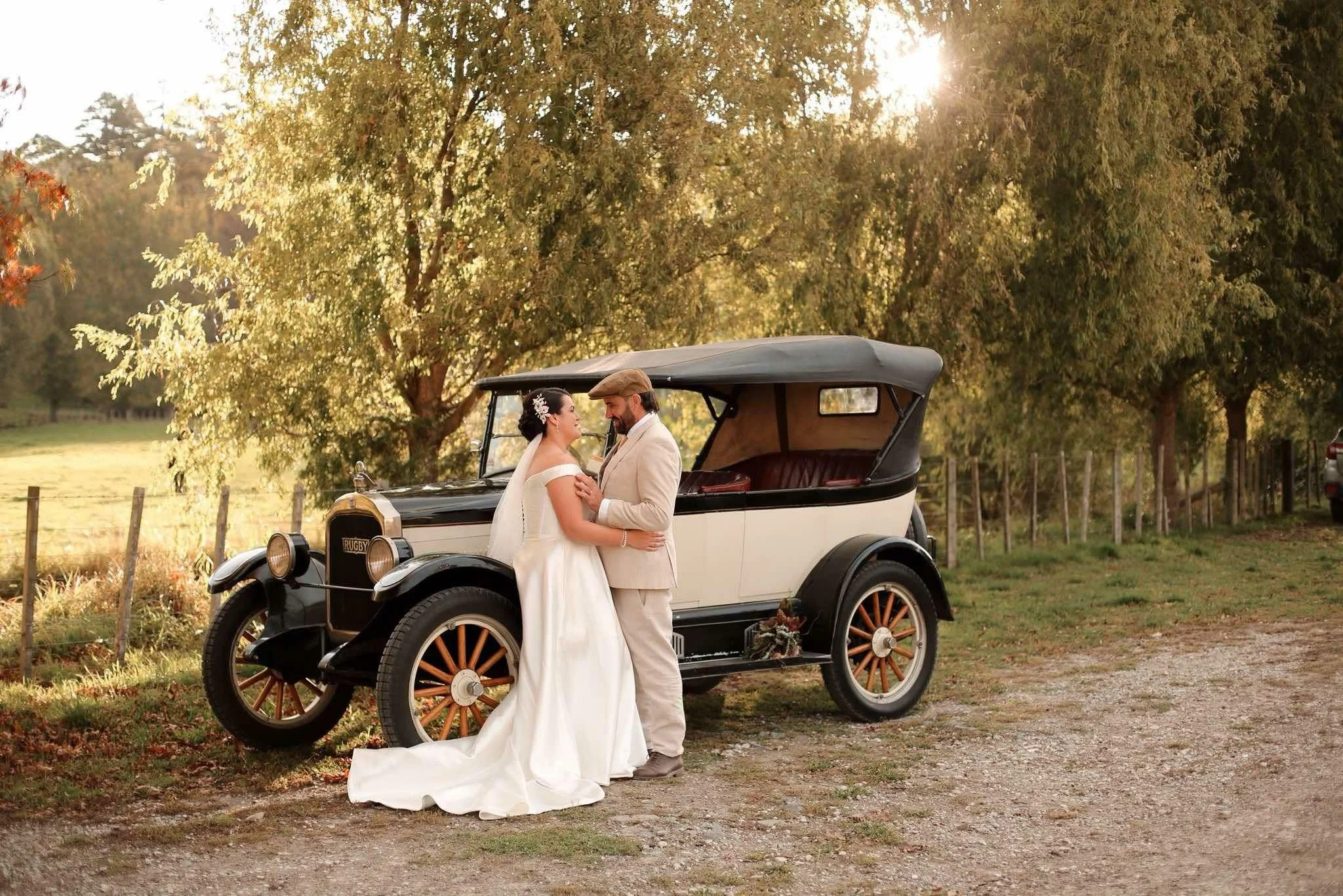 Bride and groom standing close together in front of a vintage car outdoors, with trees and sunlight in the background.
