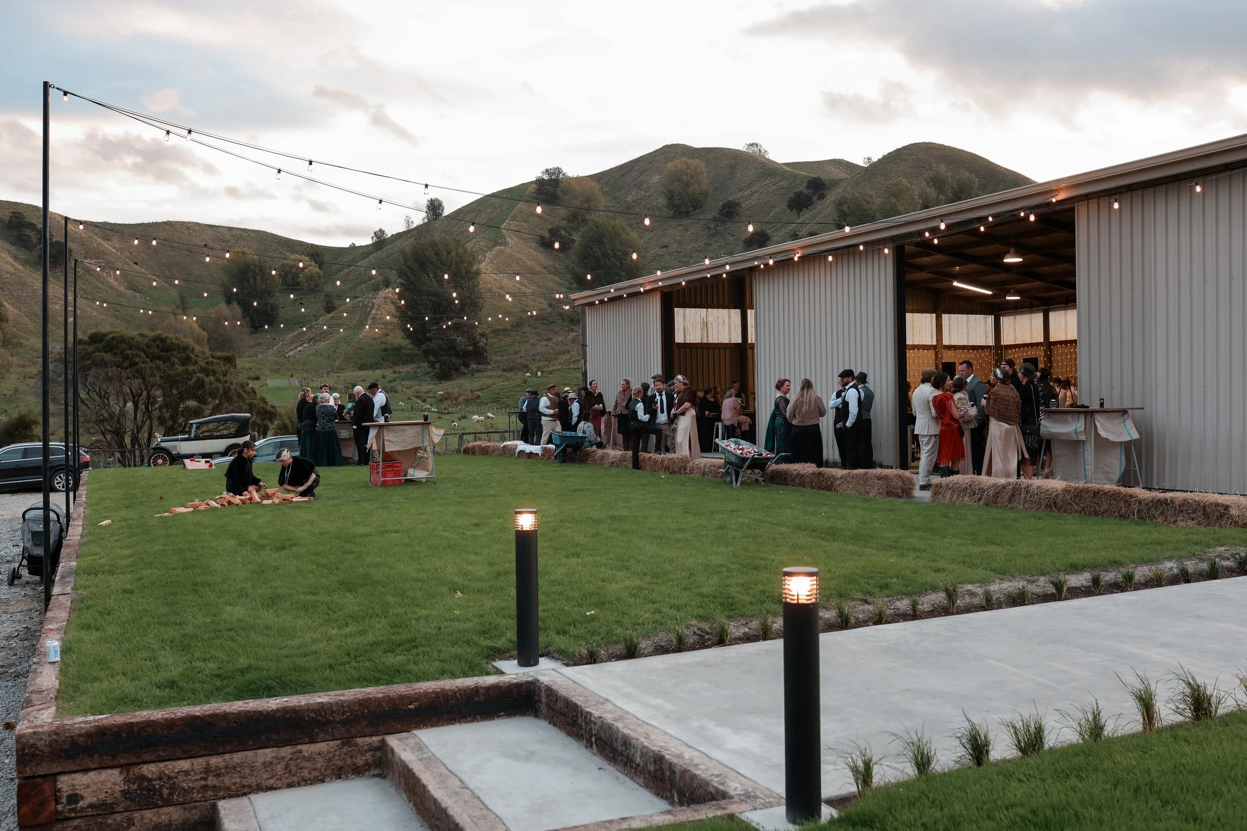 An outdoor gathering in a rural setting with a barn and string lights, people socializing, and hills in the background at dusk.