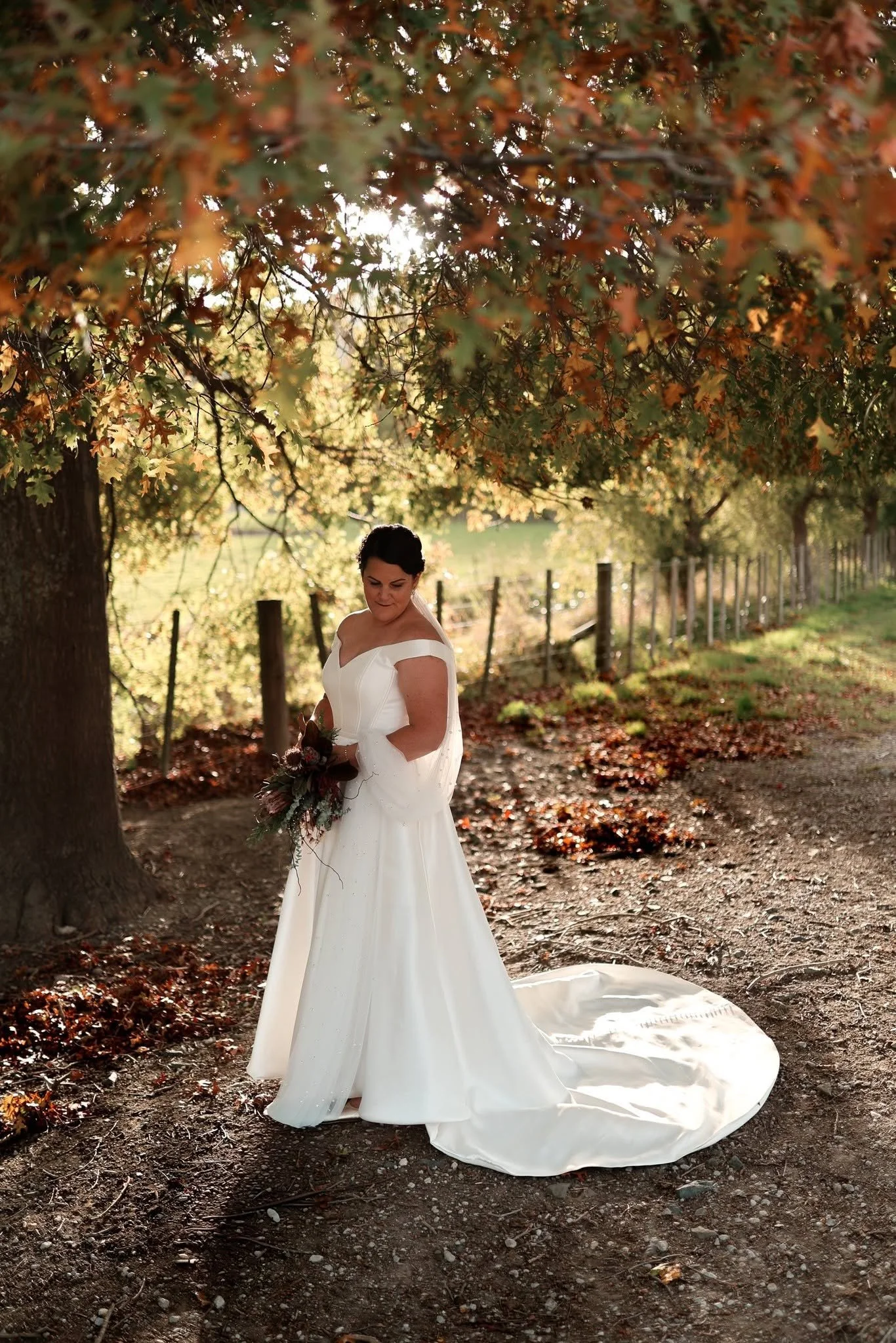 A bride in a white wedding dress holding a bouquet stands under a large tree with autumn-colored leaves, during sunset in a rural outdoor setting with a fence.