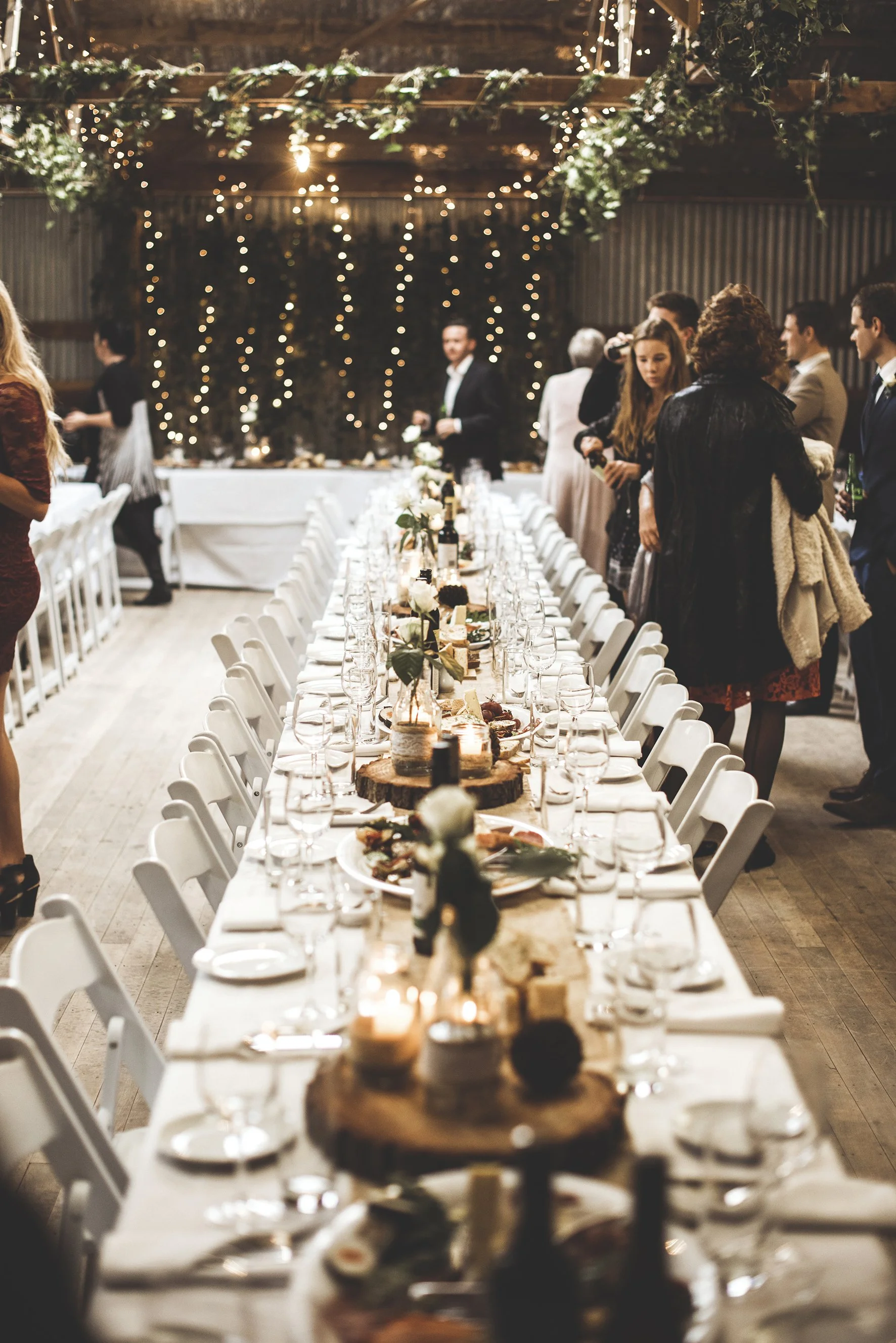 A long banquet table set for a celebration with white chairs, floral centerpieces, candles, and wine bottles. People are standing around the table, talking, in a rustic venue decorated with string lights and greenery.