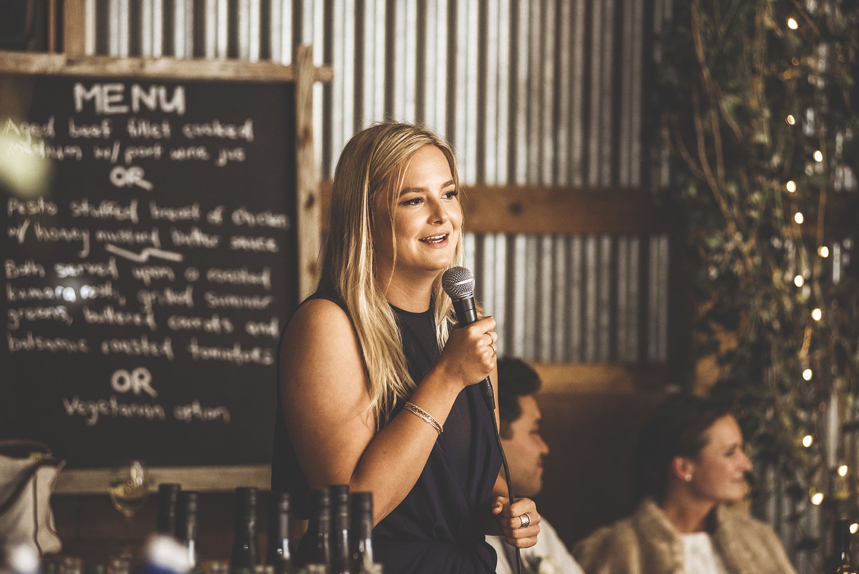 A woman with long blonde hair holding a microphone, speaking at an indoor event with a rustic background and a menu board behind her.