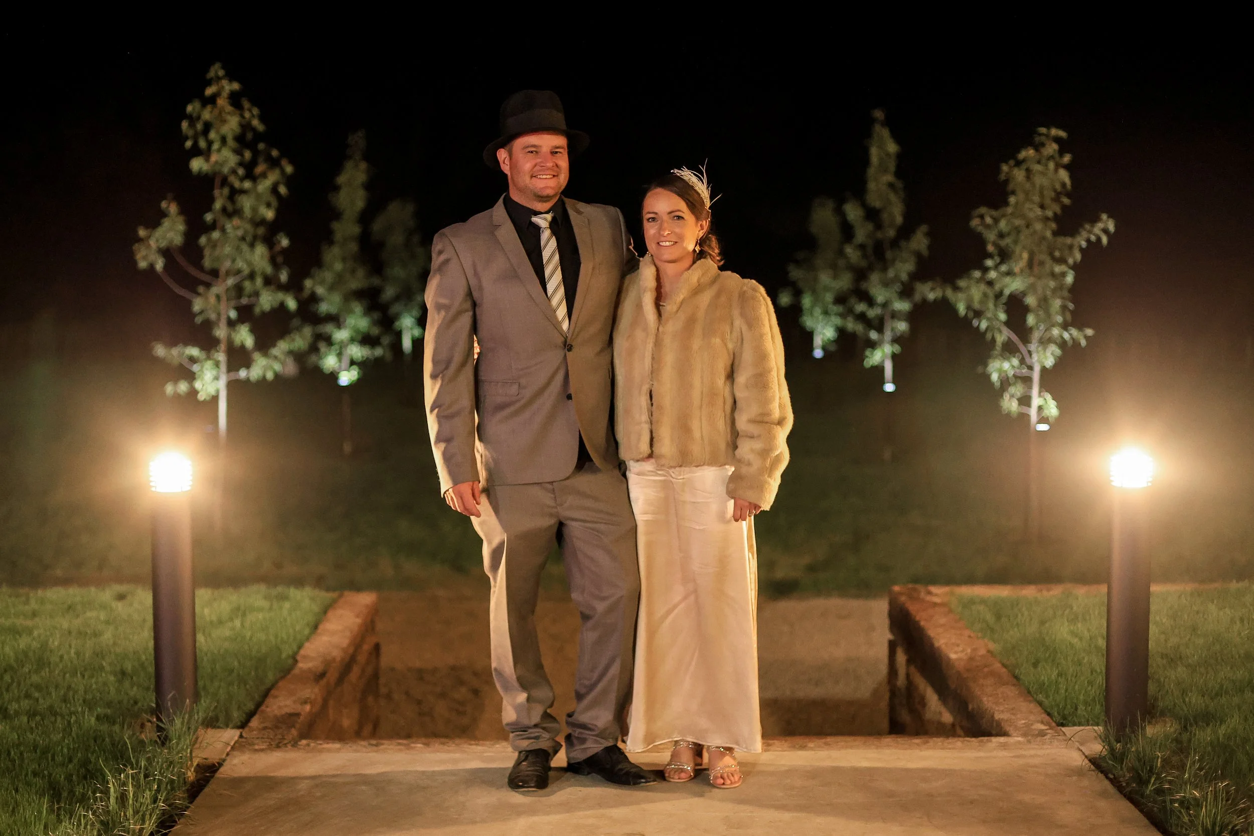 A man and woman standing together outdoors at night, dressed formally, with decorative lighting and trees in the background.