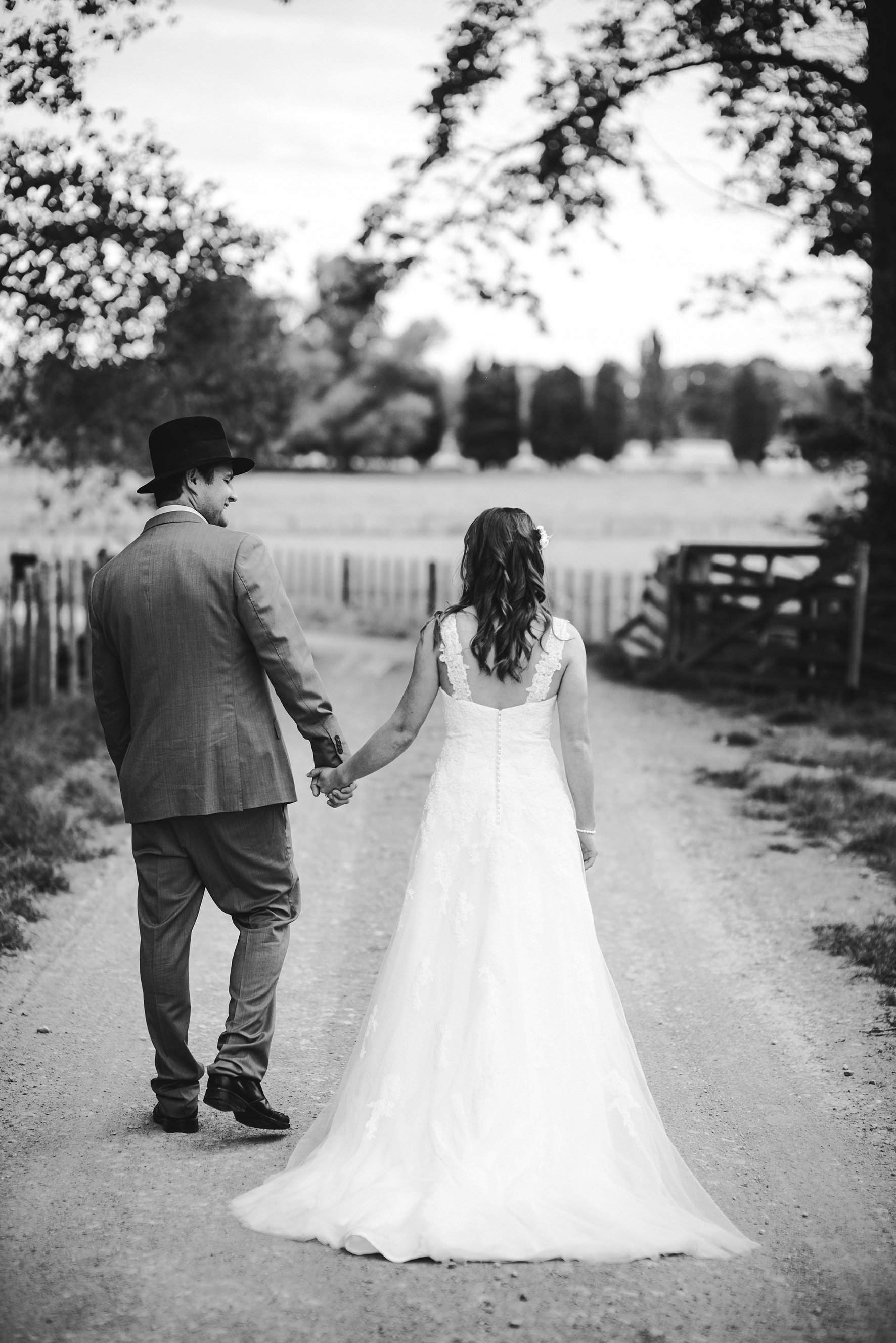 A bride and groom holding hands and walking away in a park-like setting with trees and a bench, black and white photograph