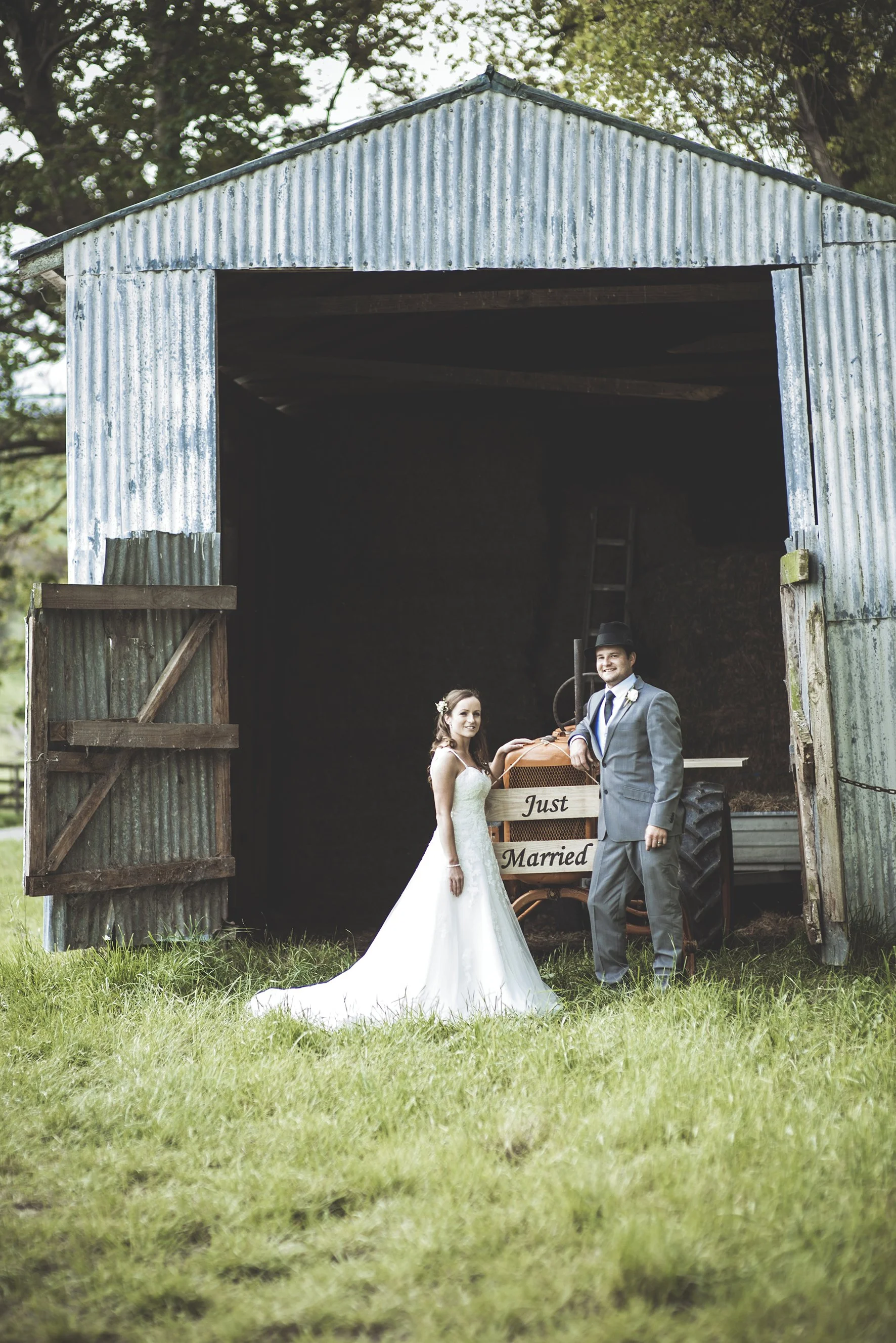 A bride and groom standing outside a rustic metal barn, smiling, with a small wagon that says "Just Married" behind them, green grass in the foreground, and trees in the background.