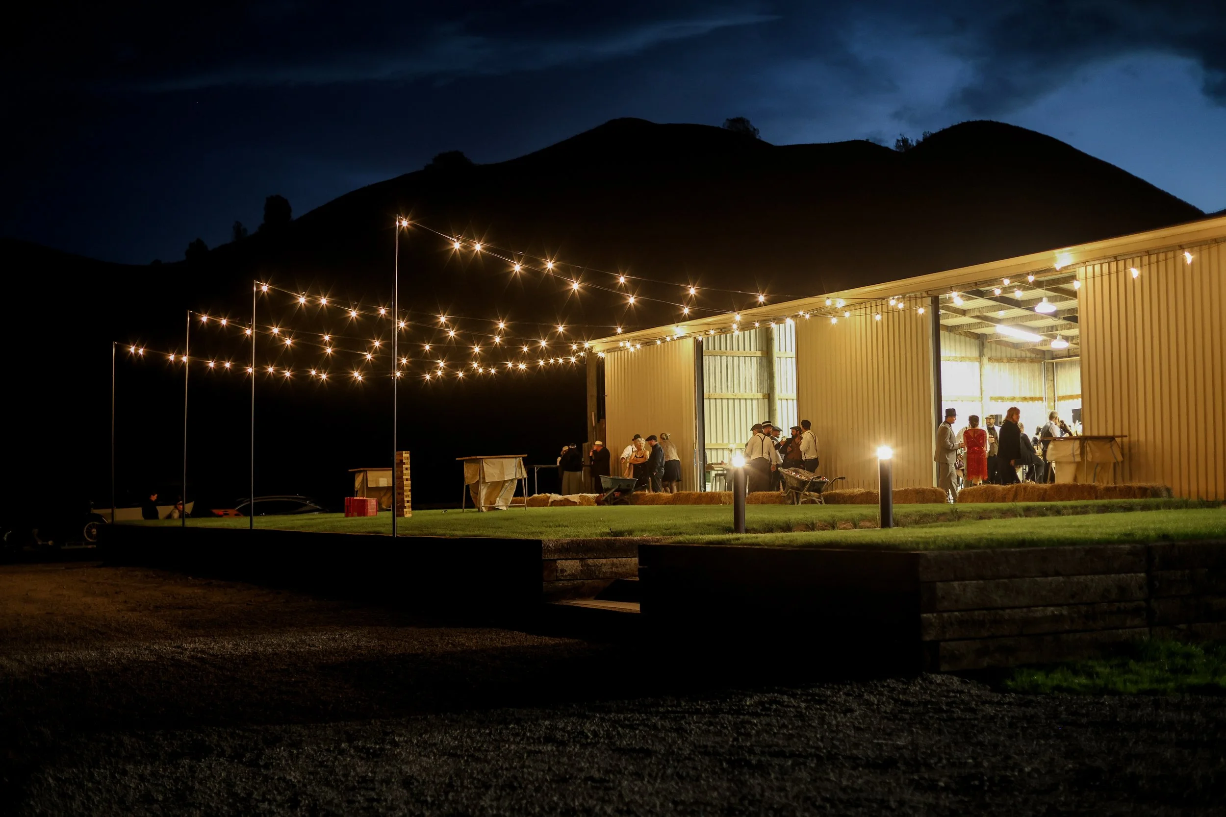 Nighttime scene of an illuminated building with people gathered inside, string lights above, and a mountain silhouette in the background.