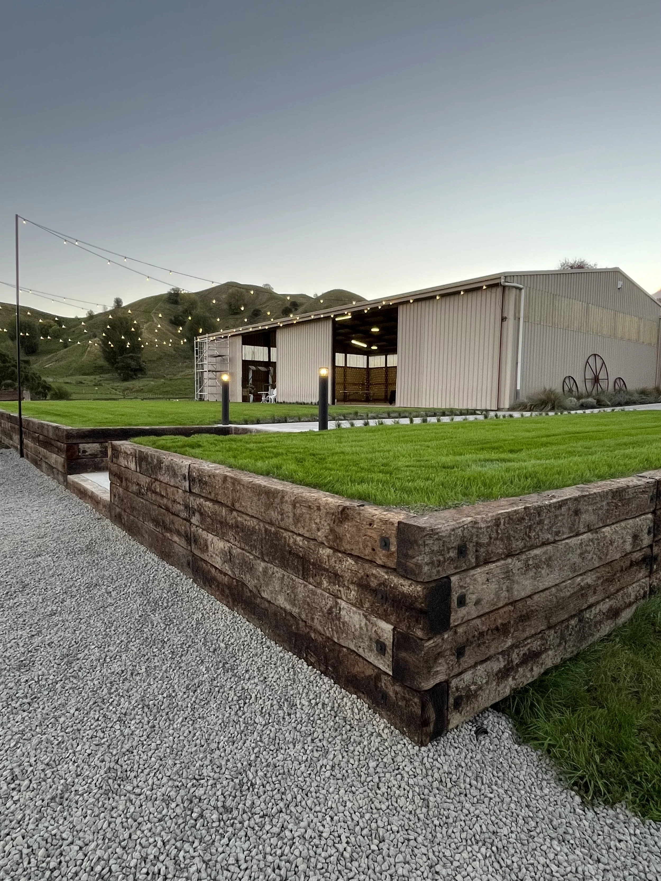 A modern barn with large wheels, string lights overhead, a small porch area with chairs, and a lush green lawn with a gravel pathway in the foreground, set against hilly landscape at dusk.