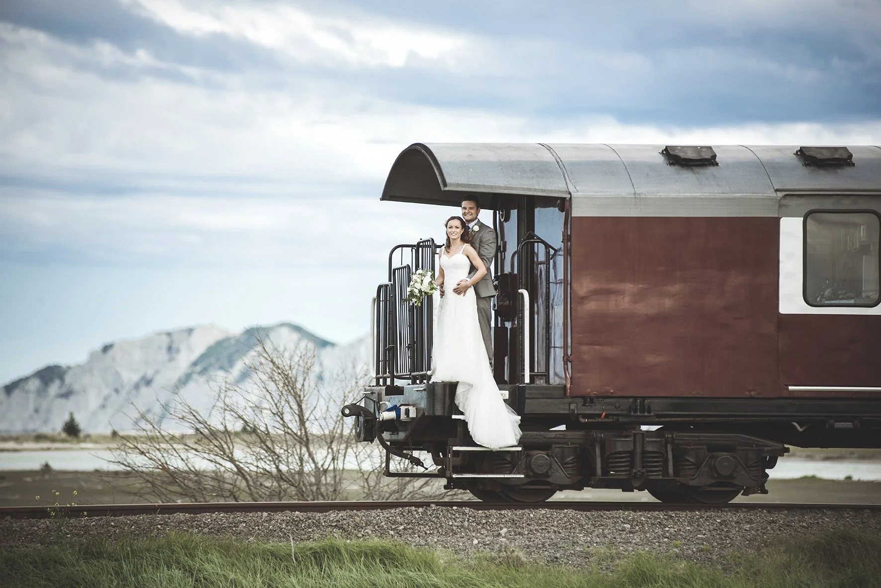 A bride and groom standing on the step of a vintage train car with mountains in the background.