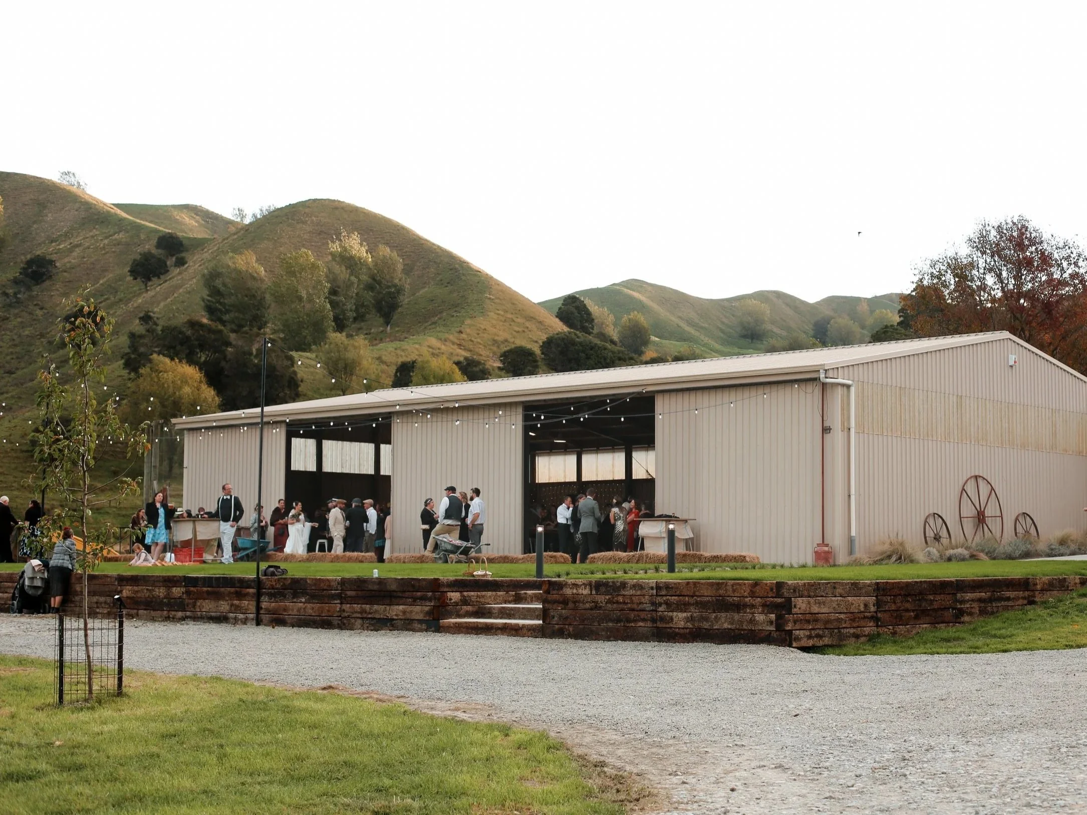 People gathered outdoors near a large industrial-style building with string lights, set against a backdrop of rolling green hills and trees, suggesting a social event or gathering.