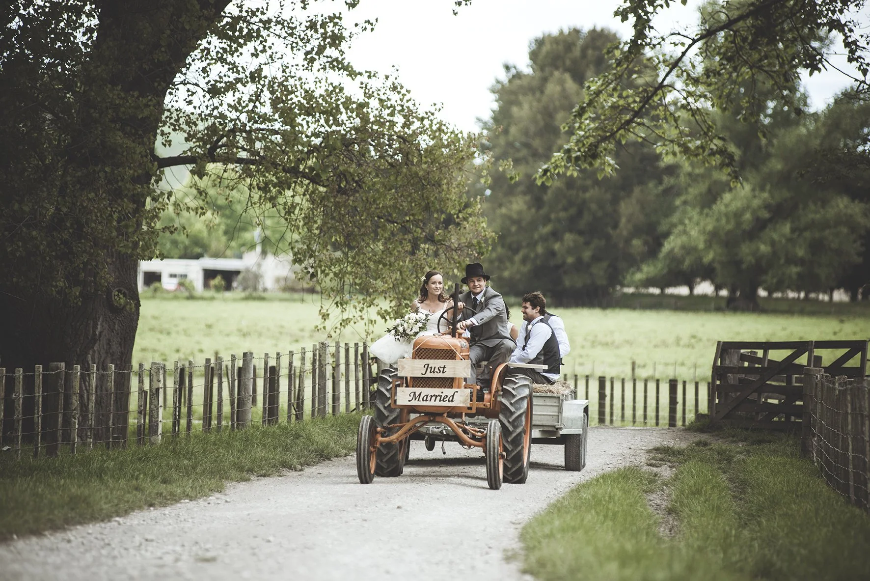 A group of people riding a tractor with a sign that reads 'Just Married,' in a rural setting with trees and a fence.