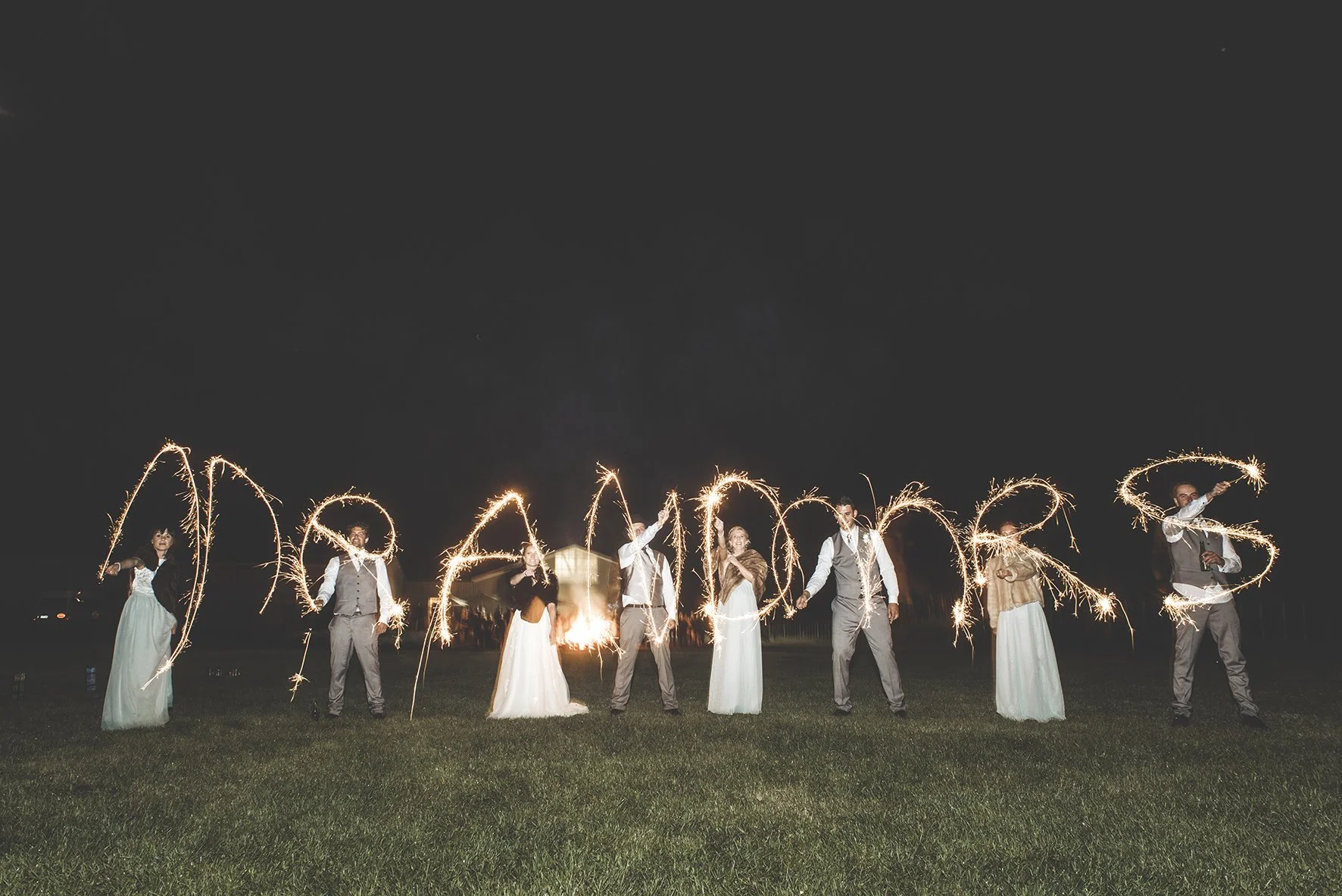 People dressed in formal attire holding sparklers in night-time outdoor celebration, spelling out the word 'DREAM' with light trails