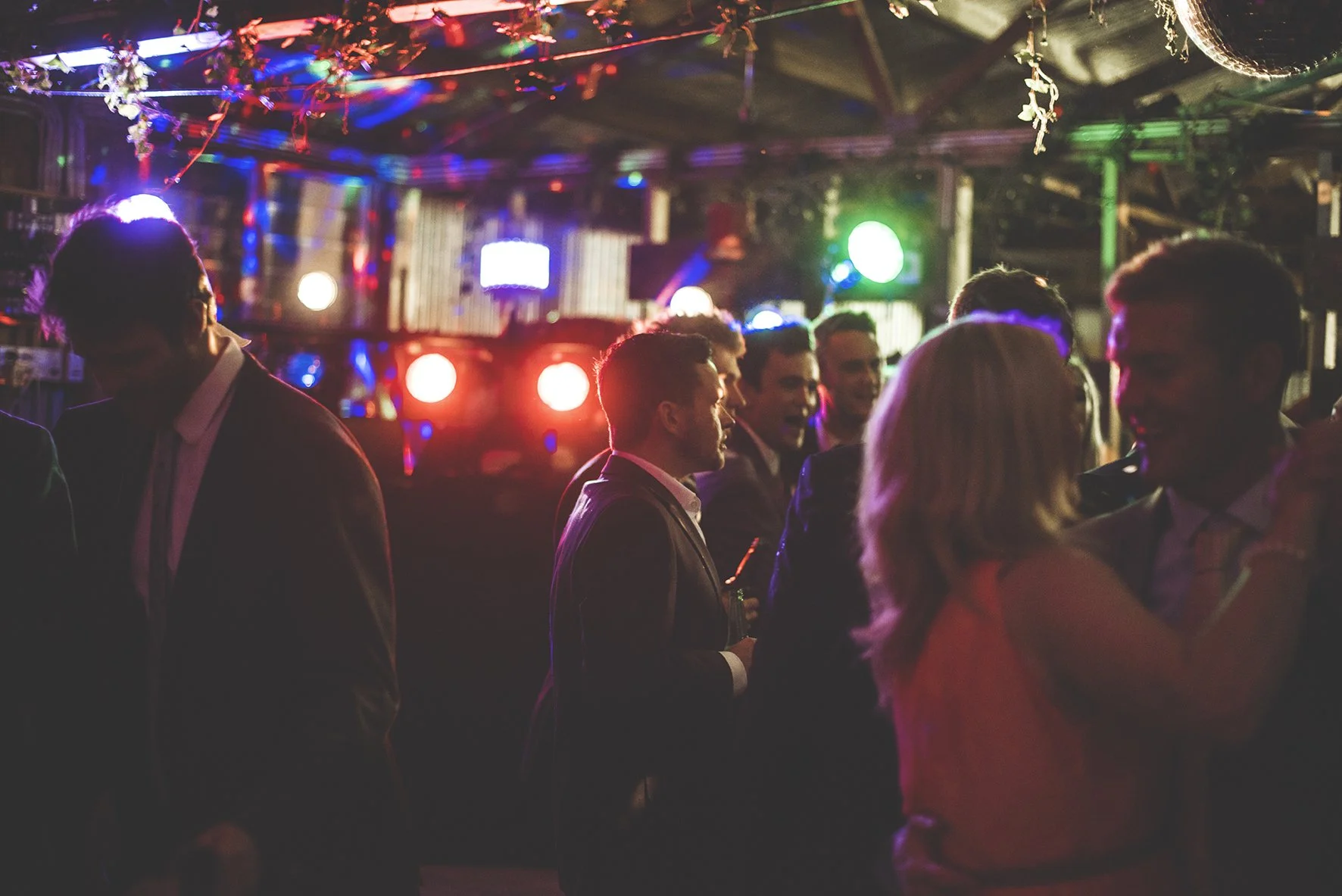 People dancing and socializing at a dimly lit party with colorful lights and a DJ setup in the background.