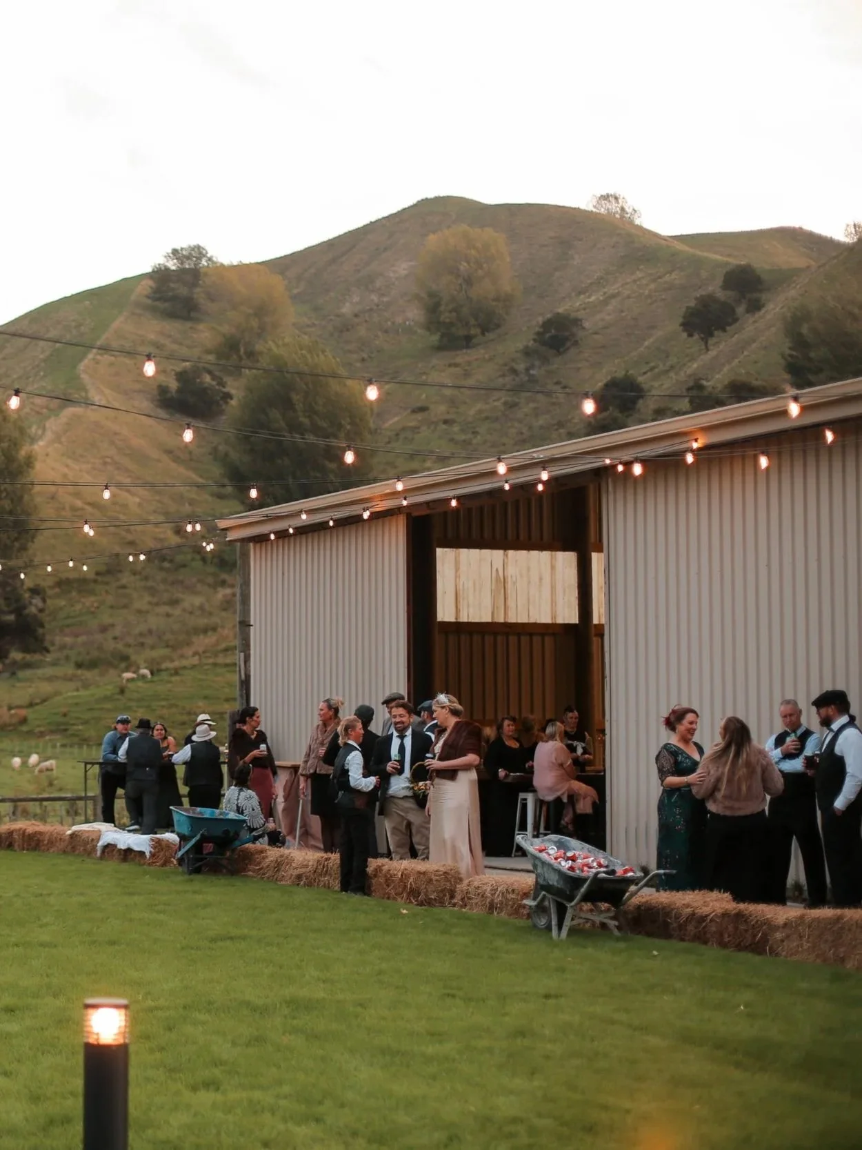 Outdoor gathering at a rustic venue with string lights, people socializing and enjoying drinks, set against a backdrop of hills and trees.