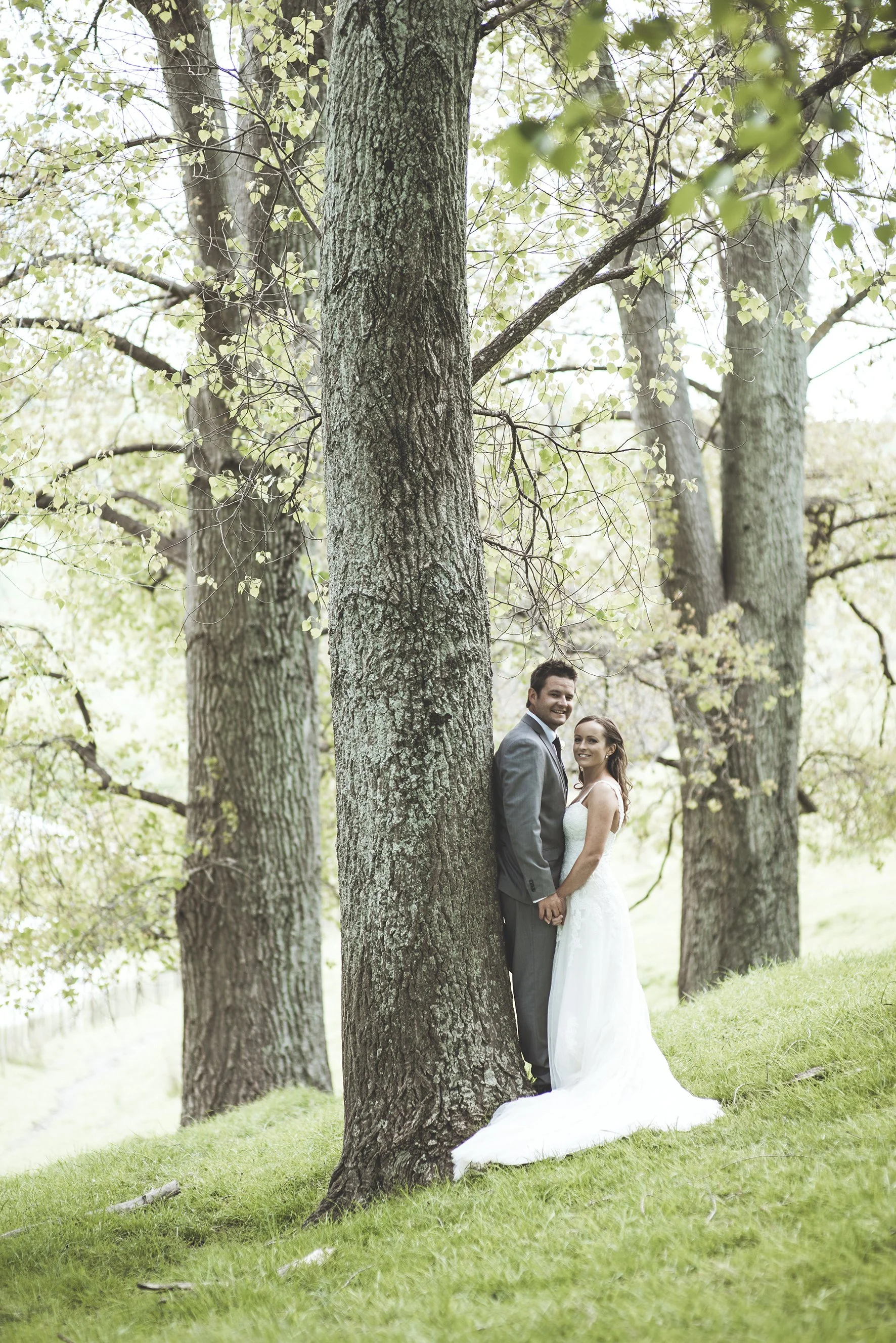 A bride and groom standing in a wooded area, holding hands, with the groom in a gray suit and the bride in a white wedding dress, partially obscured by a tree in the foreground.