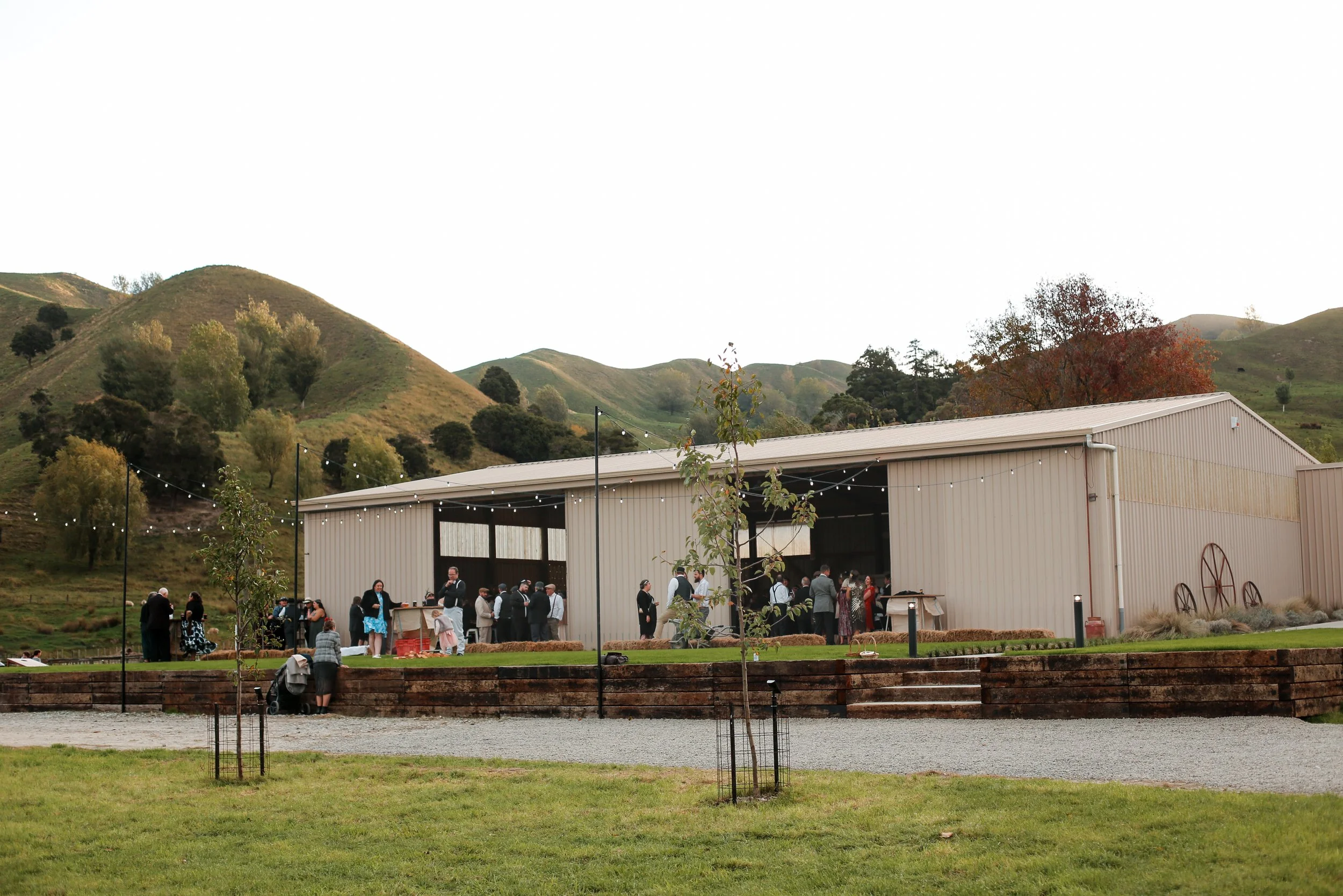 People gathered outside a large warehouse-style building with string lights, set against rolling hills with sparse trees and some autumn foliage.