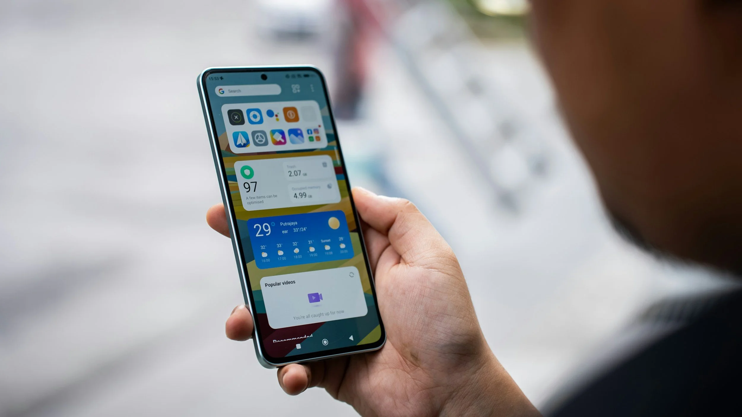 Close-up of a person holding a smartphone with a colorful home screen, weather forecast, and app icons visible.