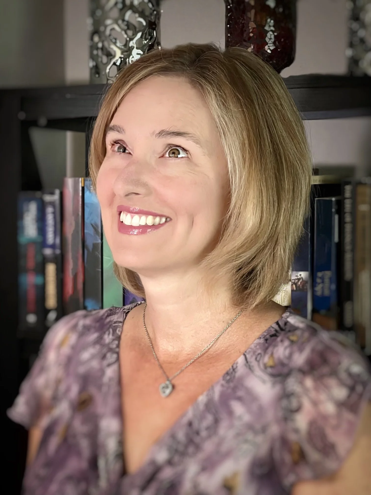 A smiling middle-aged woman with short, light brown hair is standing in front of a bookshelf filled with books. She is wearing a patterned blouse and a pendant necklace.