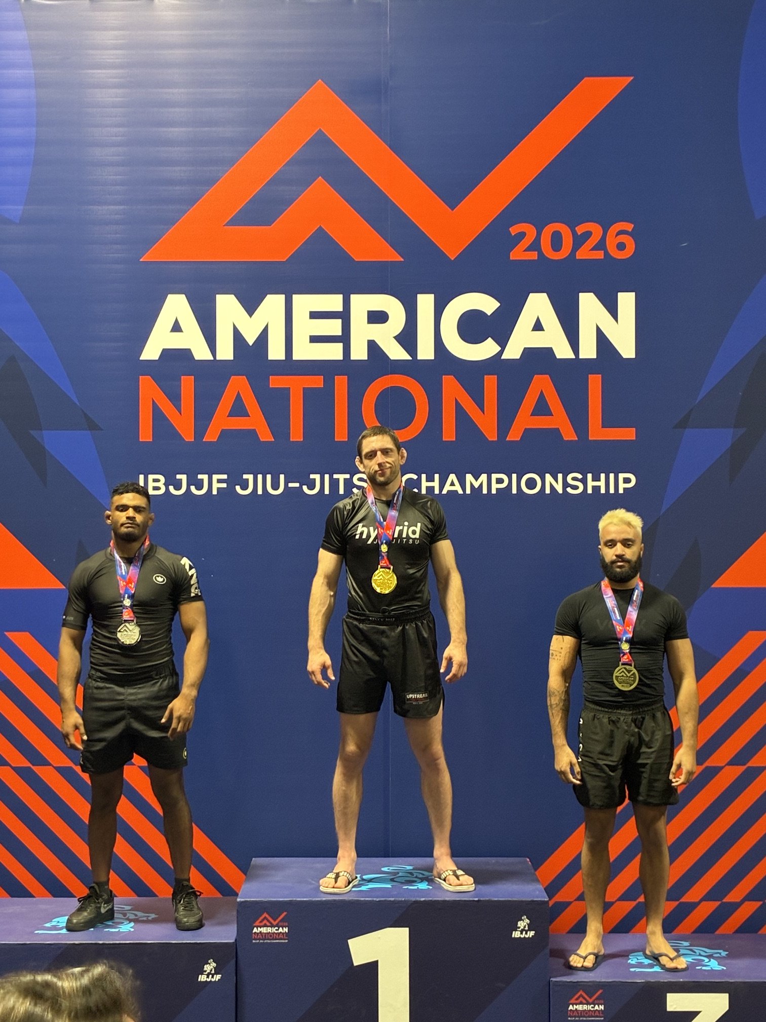 Three male athletes with medals standing on a winners' podium at the 2026 American National IBJJF Jiu-Jitsu Championship. The center athlete is the first-place winner, taller and wearing flip-flops. The athletes on the left and right are in black shorts and shirts, the left with black sneakers and the right with flip-flops. The backdrop features event branding and a large upward arrow design.