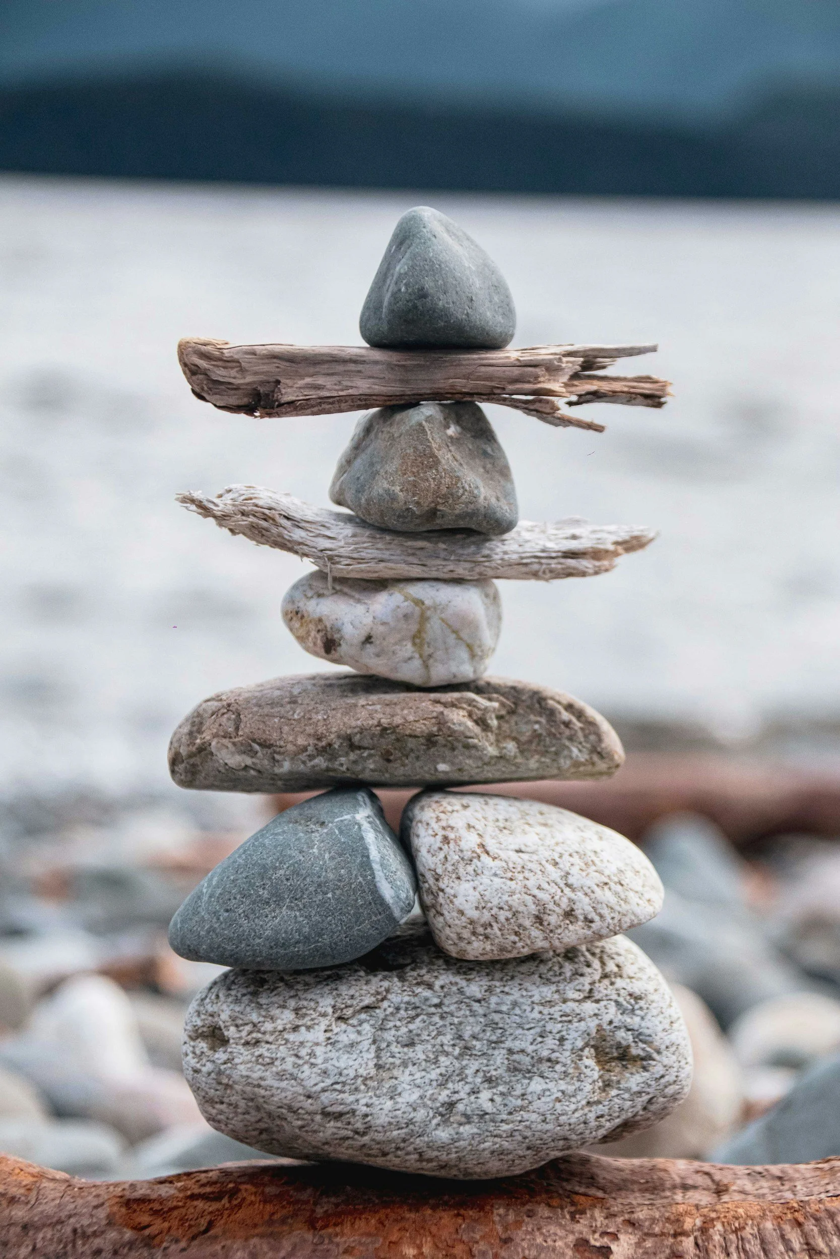 A balanced stack of nine smooth stones on a piece of driftwood at the beach, with a blurred ocean background.