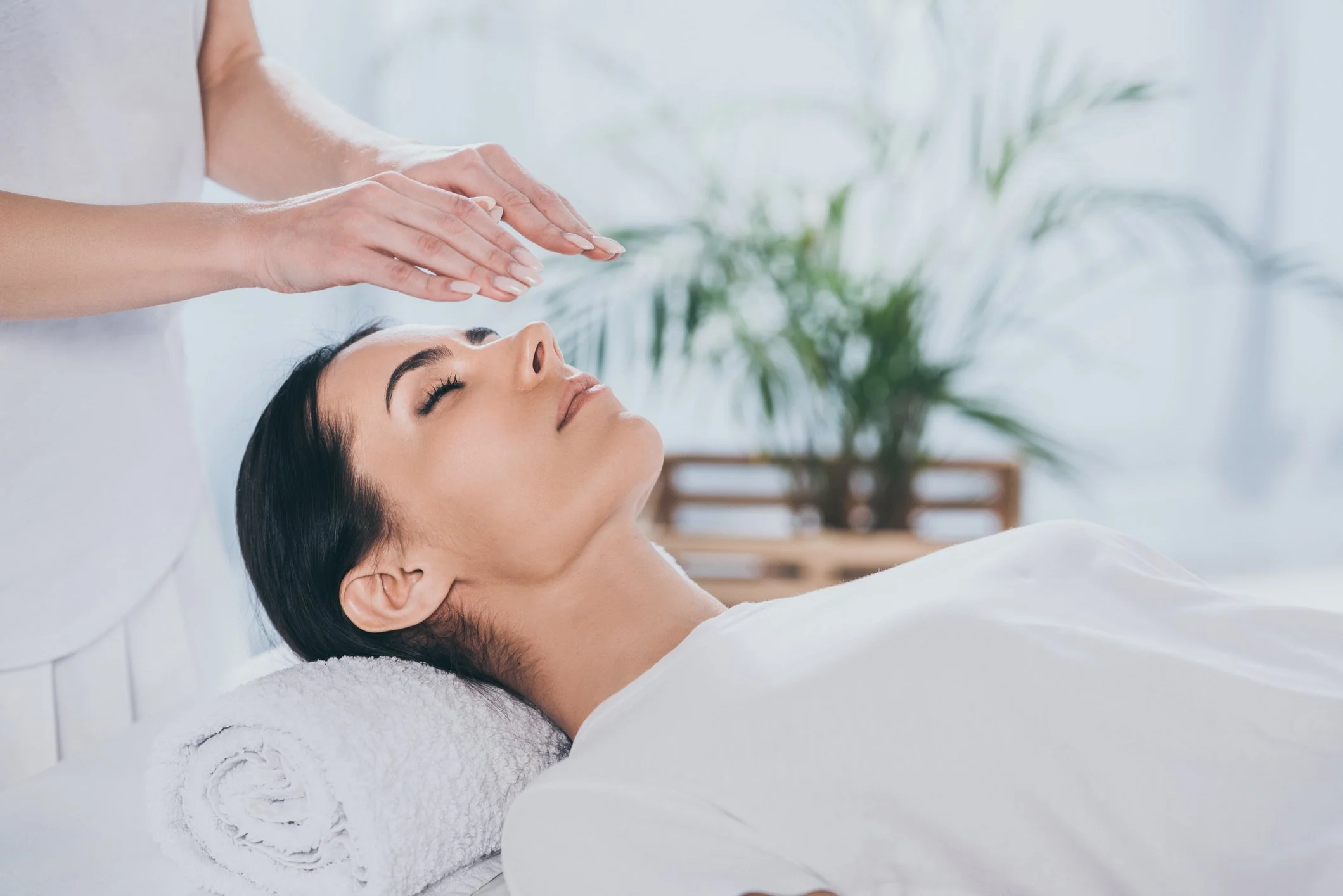 A woman with dark hair receiving a reiki treatment from a therapist in a serene spa setting, with pale blue walls and a green plant in the background.