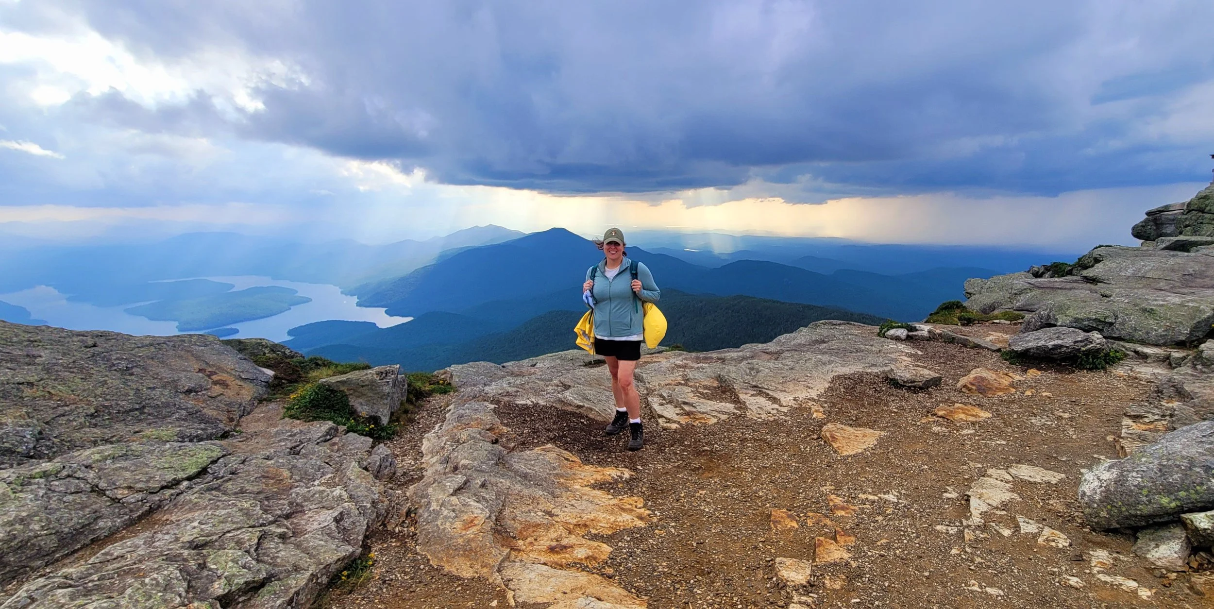 A woman hiking on rocky terrain on a mountain with a view of a lake and mountains under a cloudy sky with some sunlight breaking through.