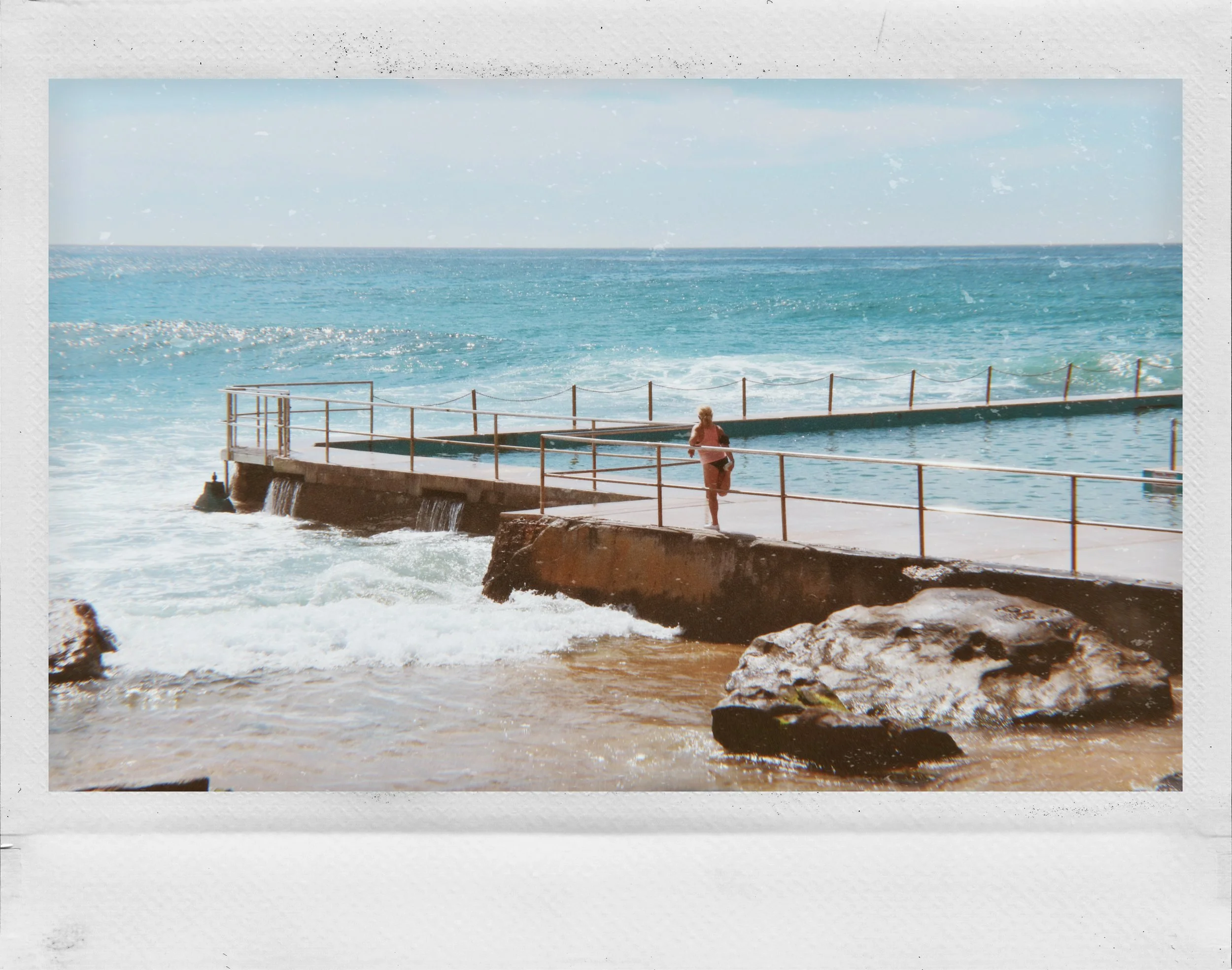 A person standing on a concrete pier with a metal railing, looking out at the ocean with blue waves and a partly cloudy sky.
