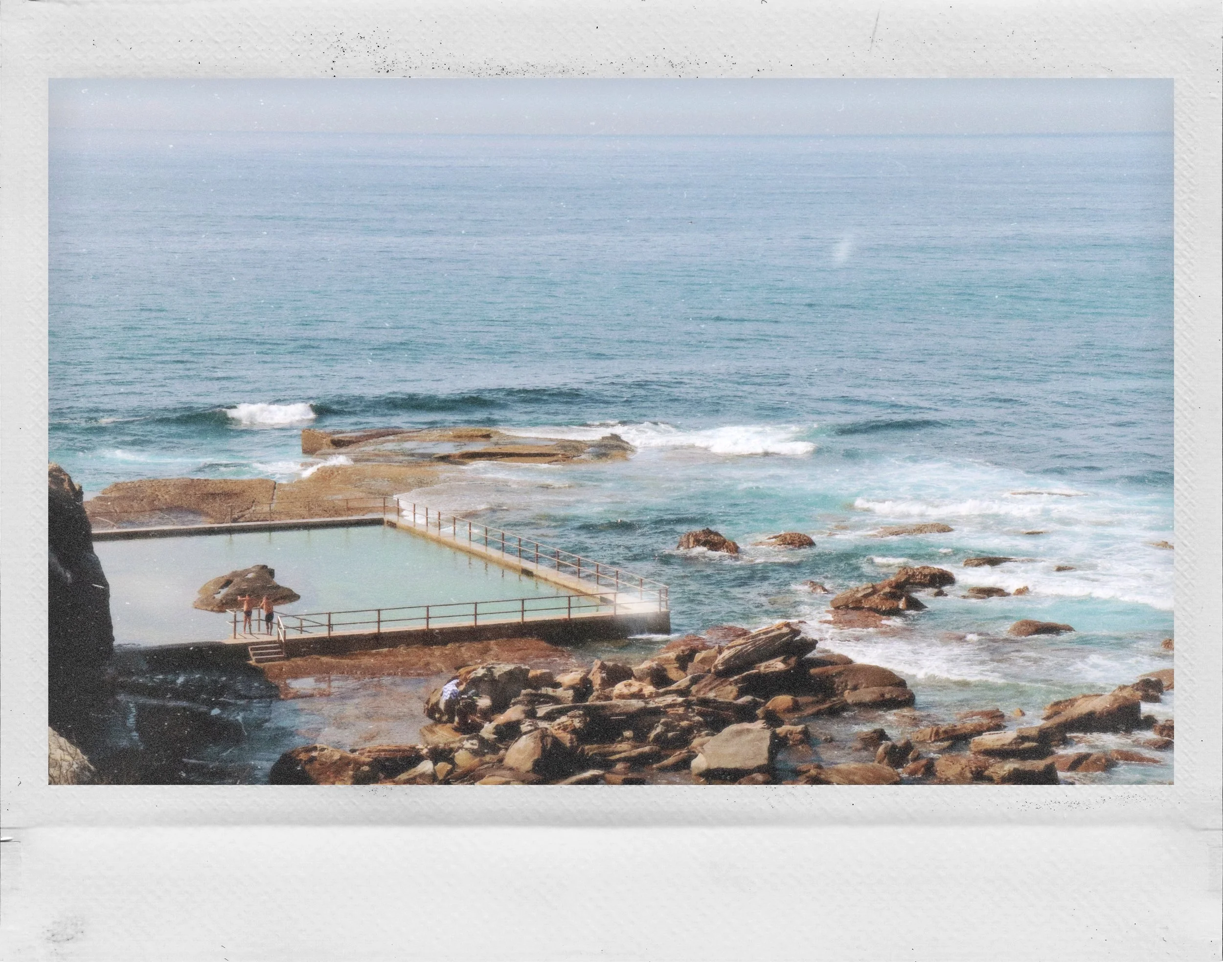 Seaside scene with rocky shoreline, ocean waves, and a pool with people on the lower left.