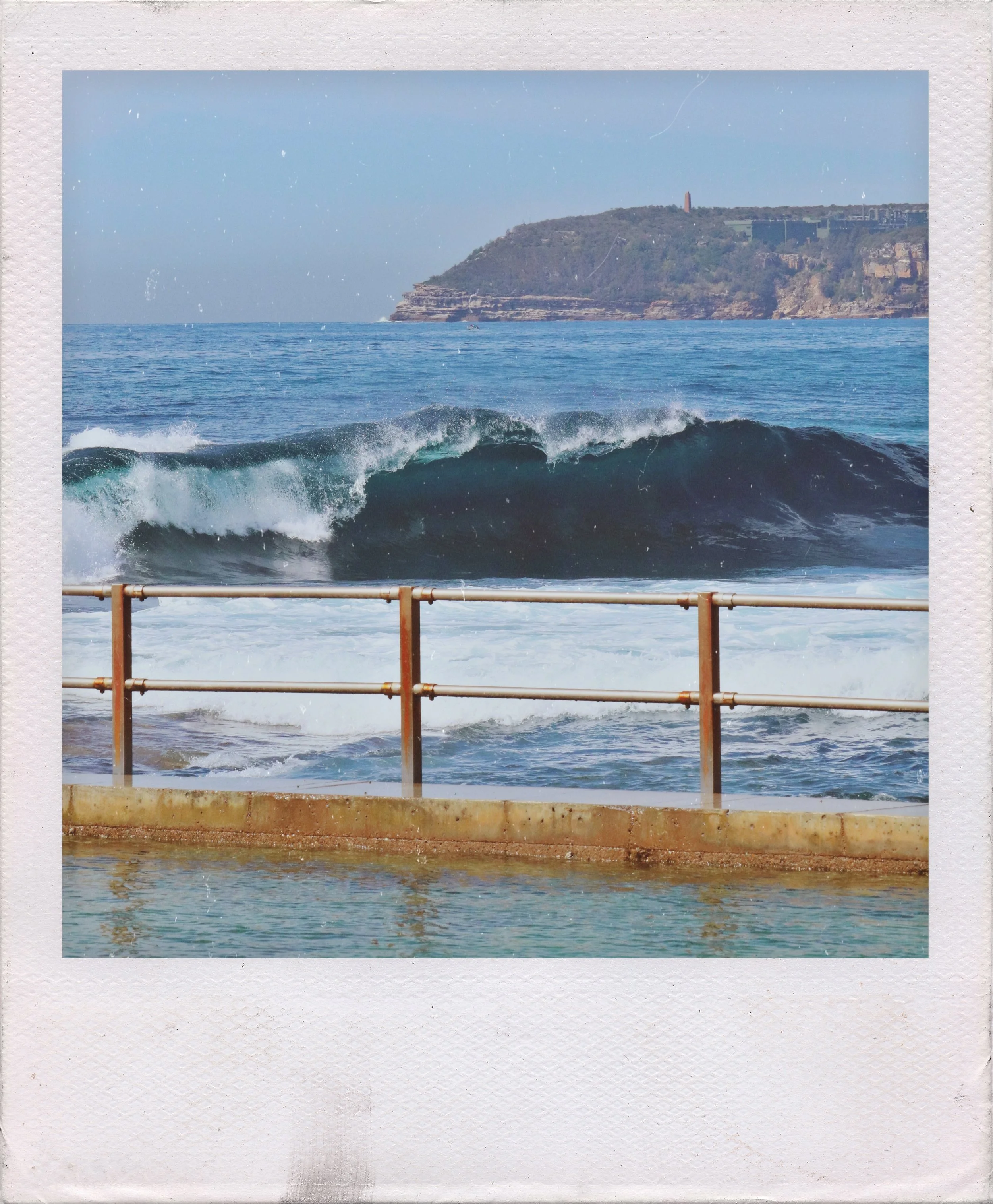 Ocean waves crashing against a barrier with a headland and cliffs in the background.