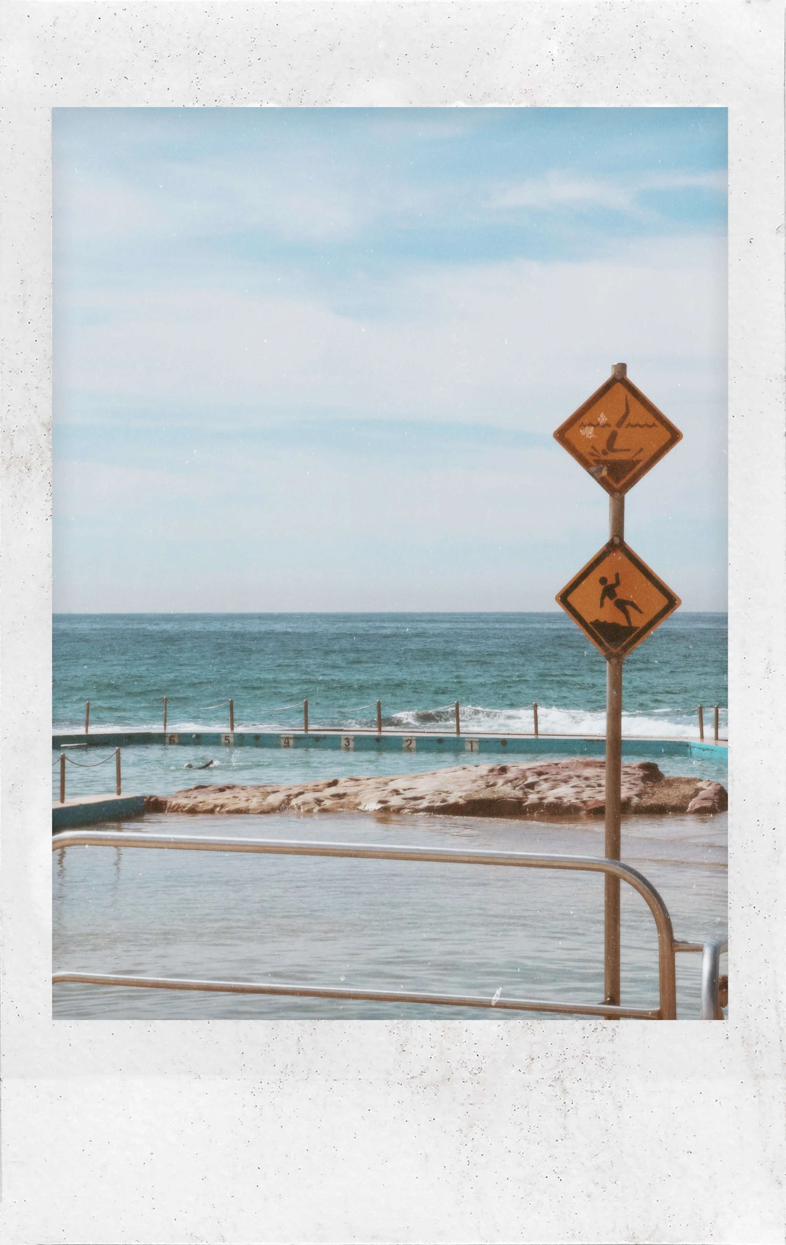 Beach scene with ocean, sky, swimming pool, and two yellow warning signs about slippery surfaces and falling rocks.