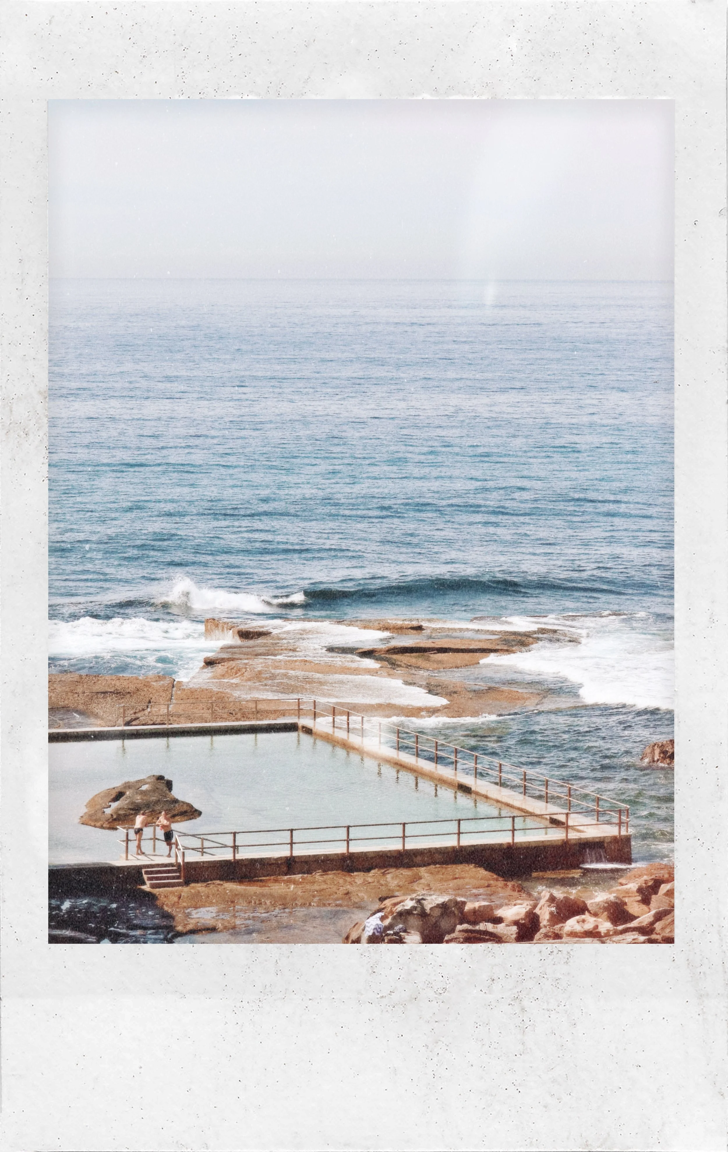 Ocean view with waves crashing on rocks and a swimming pool with a railing, two people standing by the pool, and a weathered concrete wall surrounding the pool area.
