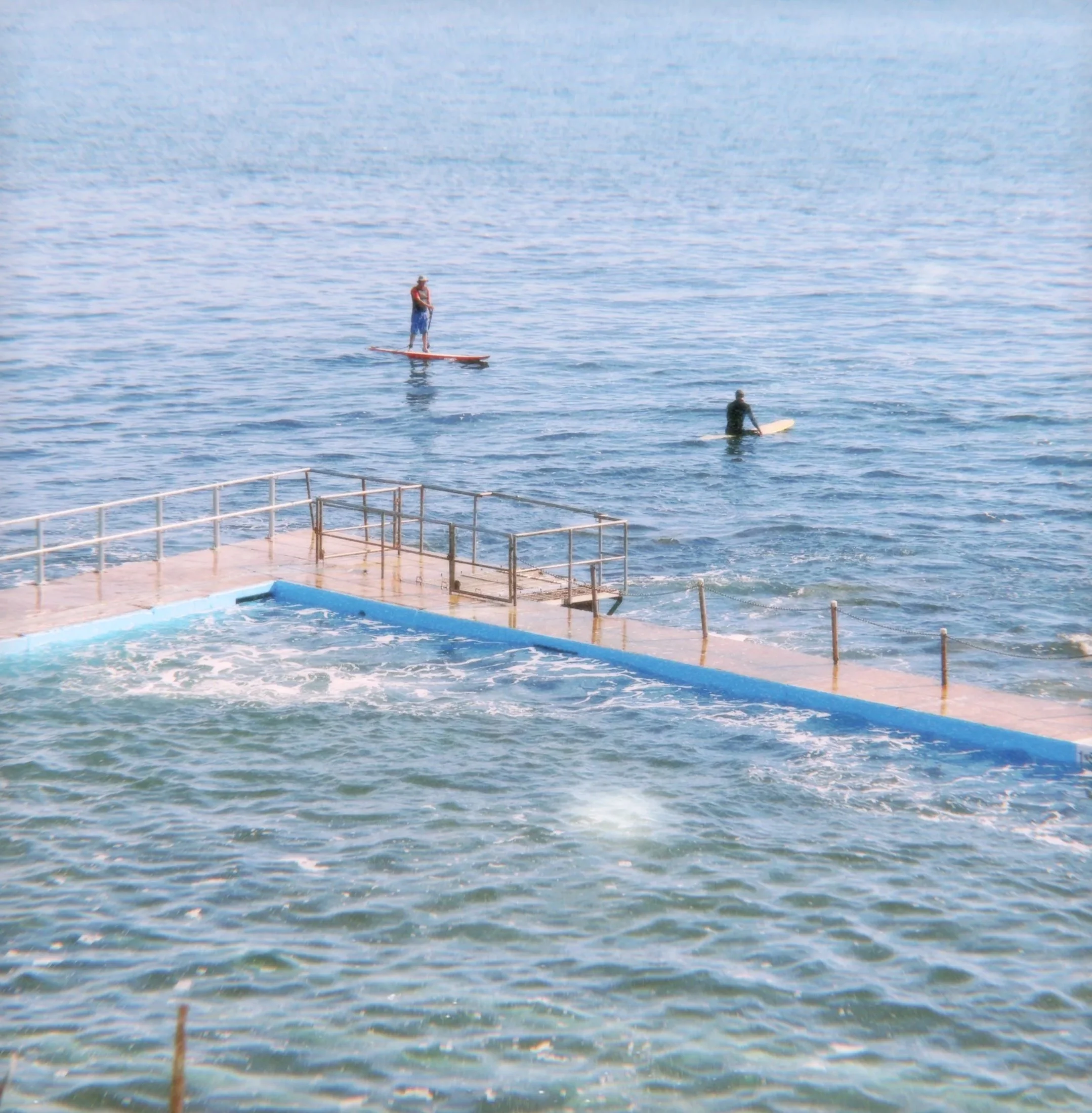 Two people stand on surfboards in a calm body of water, with the horizon and water extending into the distance.