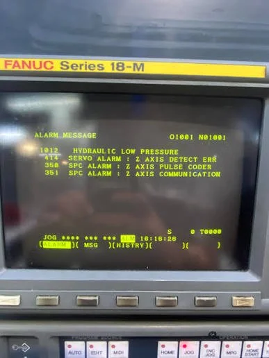 Close-up of a computer monitor displaying an alarm message and status codes, with a label indicating it's a FANUC Series 18-M CNC controller.
