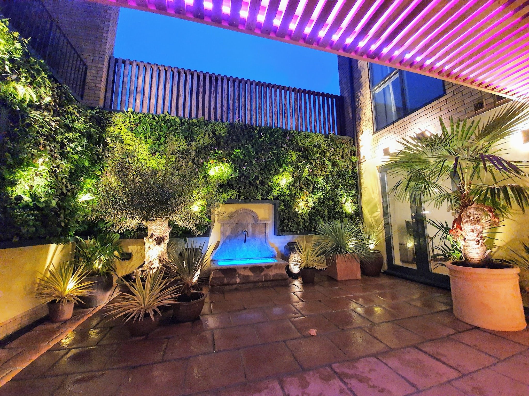 An outdoor patio area at dusk with potted plants and trees, a water fountain, brick flooring, and colorful lighting on the patio ceiling and wall.