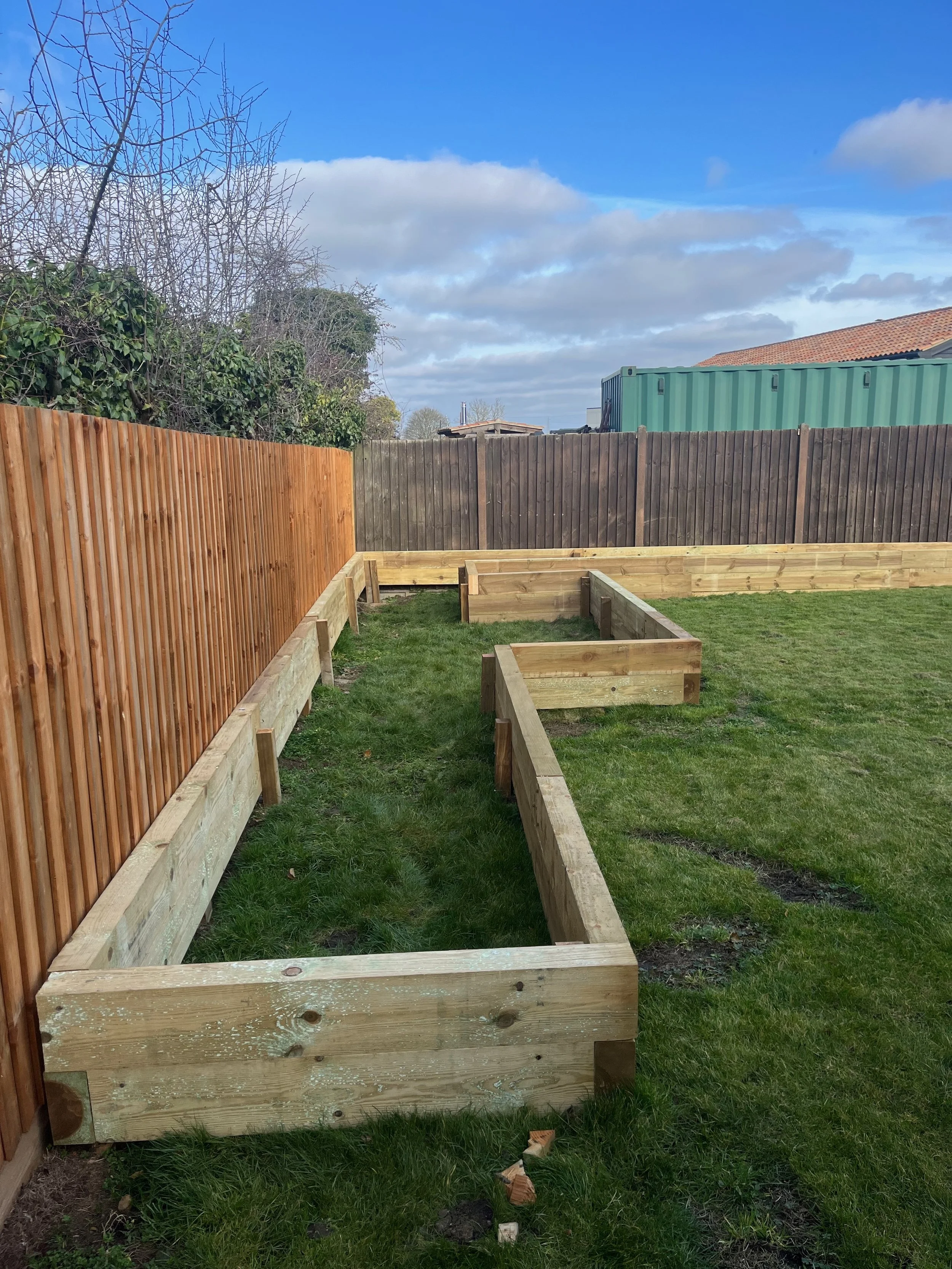A backyard garden with newly constructed wooden raised garden beds on green grass, black and brown wooden fences, and a blue sky with scattered clouds in the background.